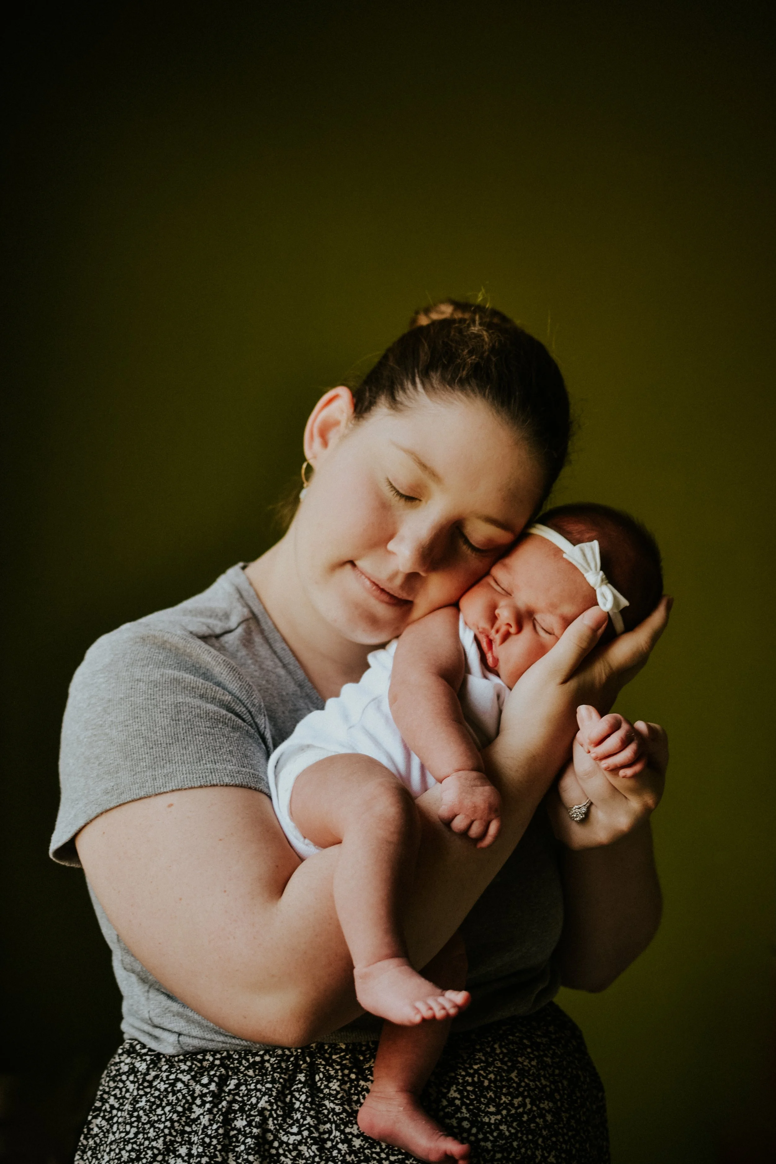 A woman with dark hair in a bun holding a newborn baby close to her face, both with their eyes closed, against a dark background.