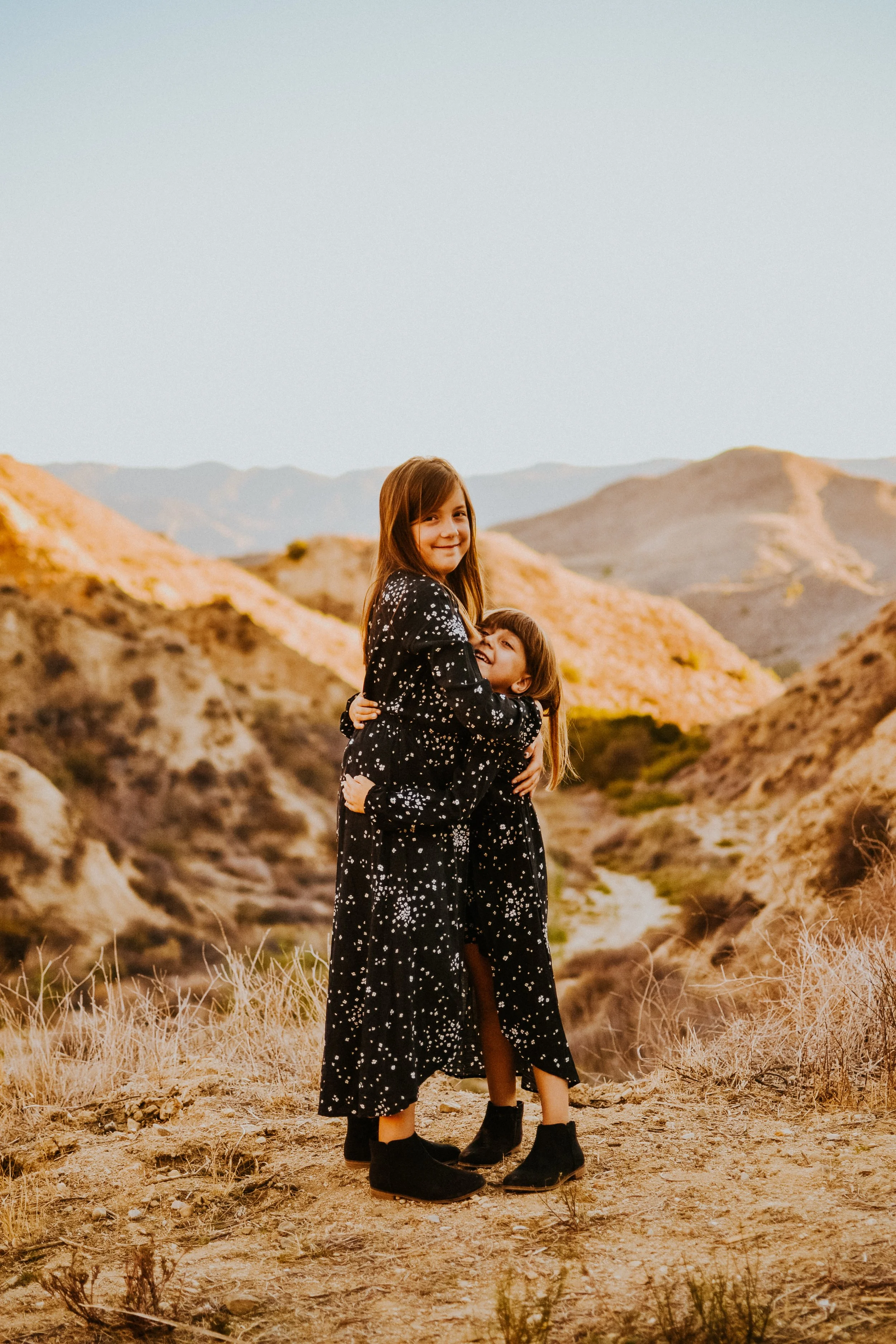 Two girls in matching black dresses with white speckled patterns hugging in a desert landscape with hills in the background.