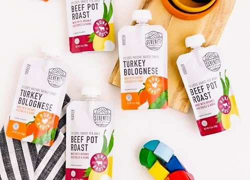 Packaged turkey bolognese and beef pot roast soup products from Serenity Foods displayed on a white surface with a cutting board and colorful utensils nearby.