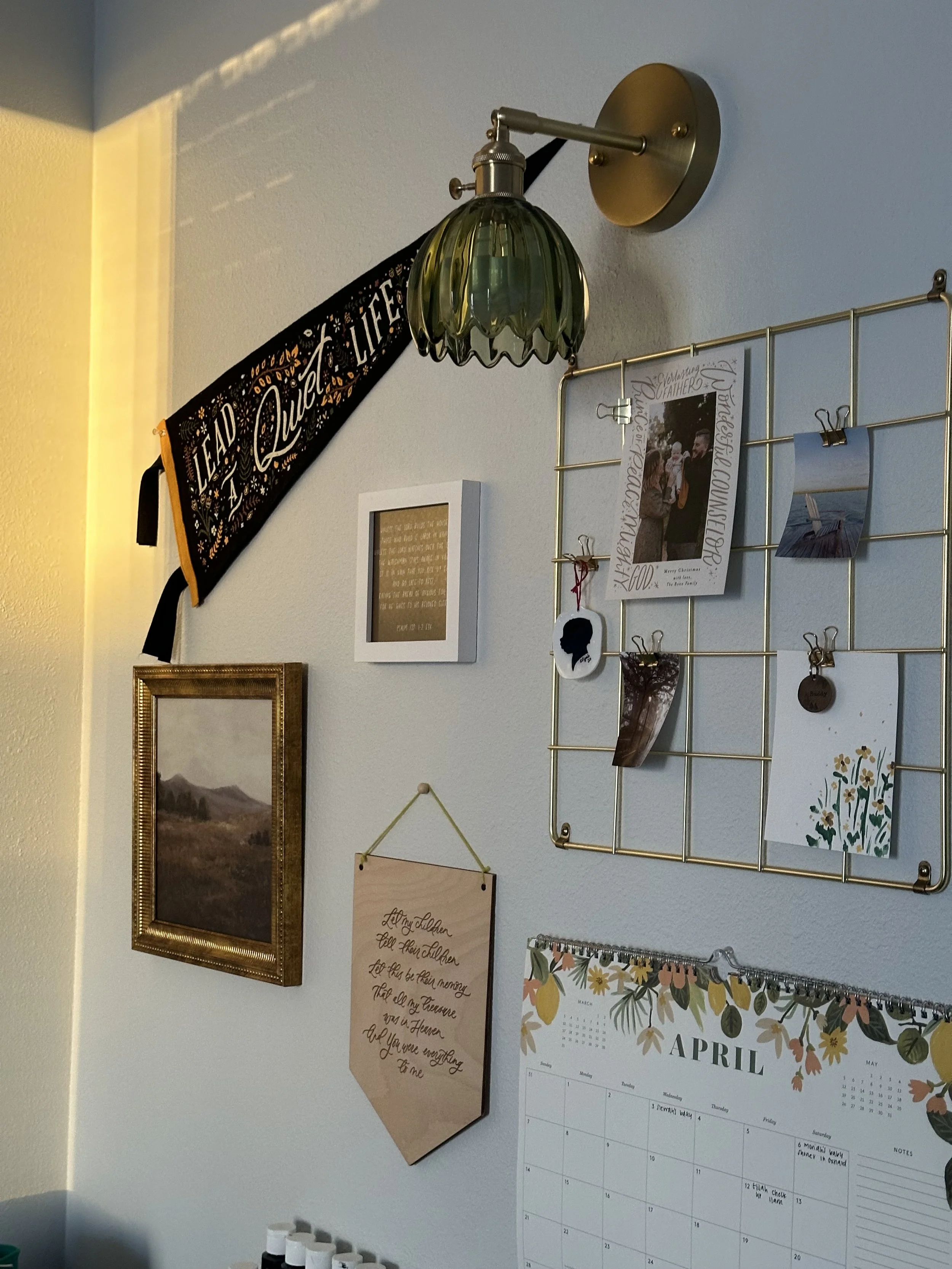 Decorative wall with photo frames, a calendar, a wall hanging with quote, a grid with photos, and a vintage-style wall sconce with a green glass shade.