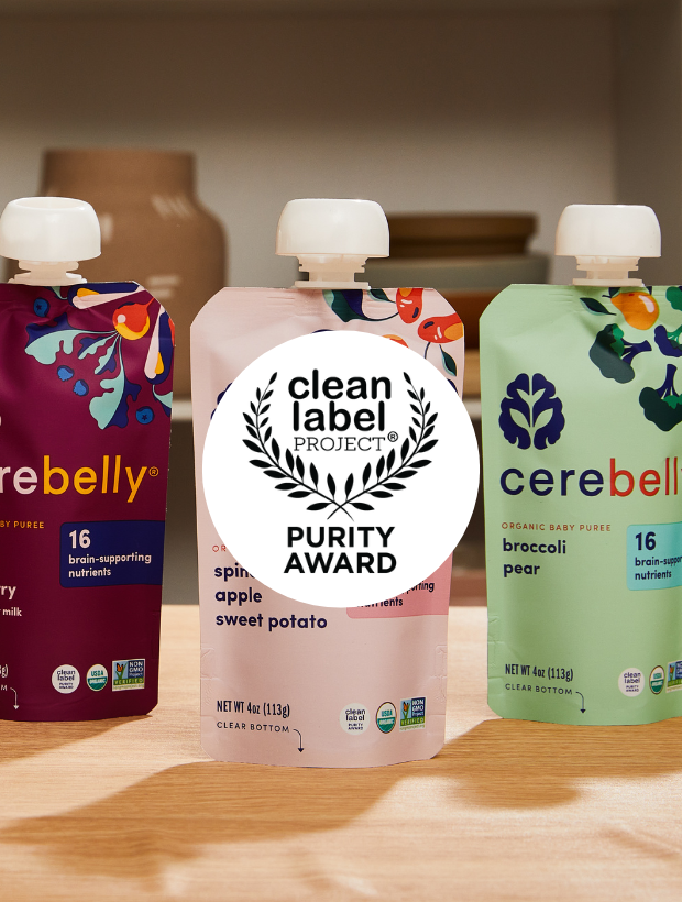 Three pouches of Cerebelly organic baby food on a wooden surface, with a large white circular Clean Label Project Purity Award sticker in the center. The flavors are cherry, apple, sweet potato on the left; spinach, apple, sweet potato in the middle; broccoli, pear on the right.