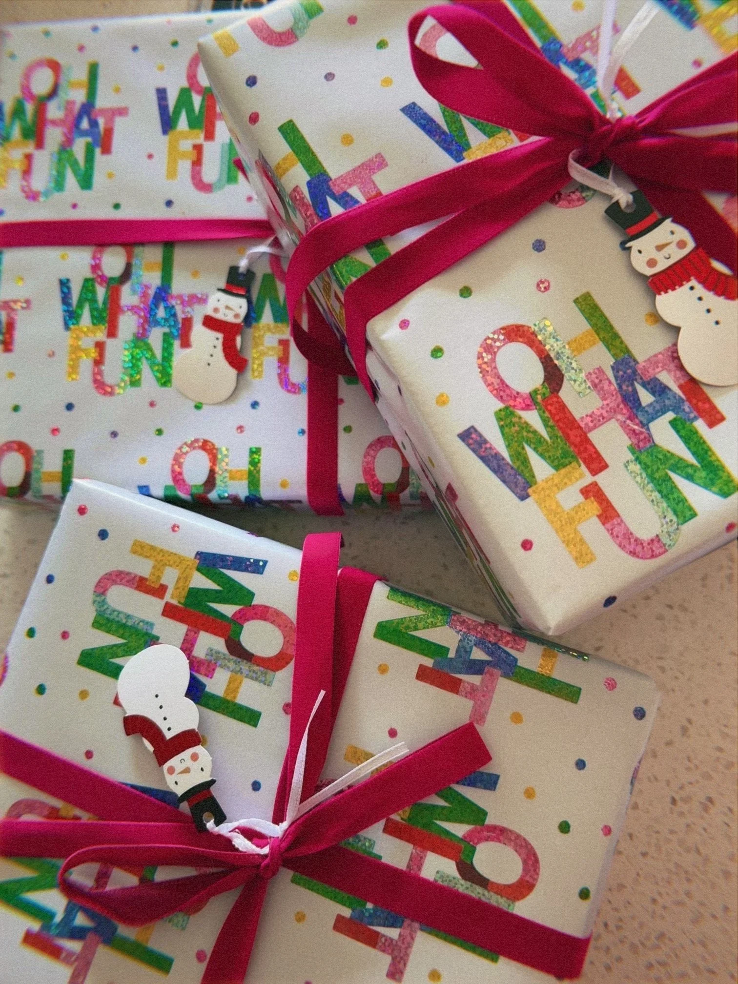 Three gift boxes wrapped in colorful, festive paper with the phrase 'Oh What Fun' and a snowman ornament attached, decorated with pink ribbons.