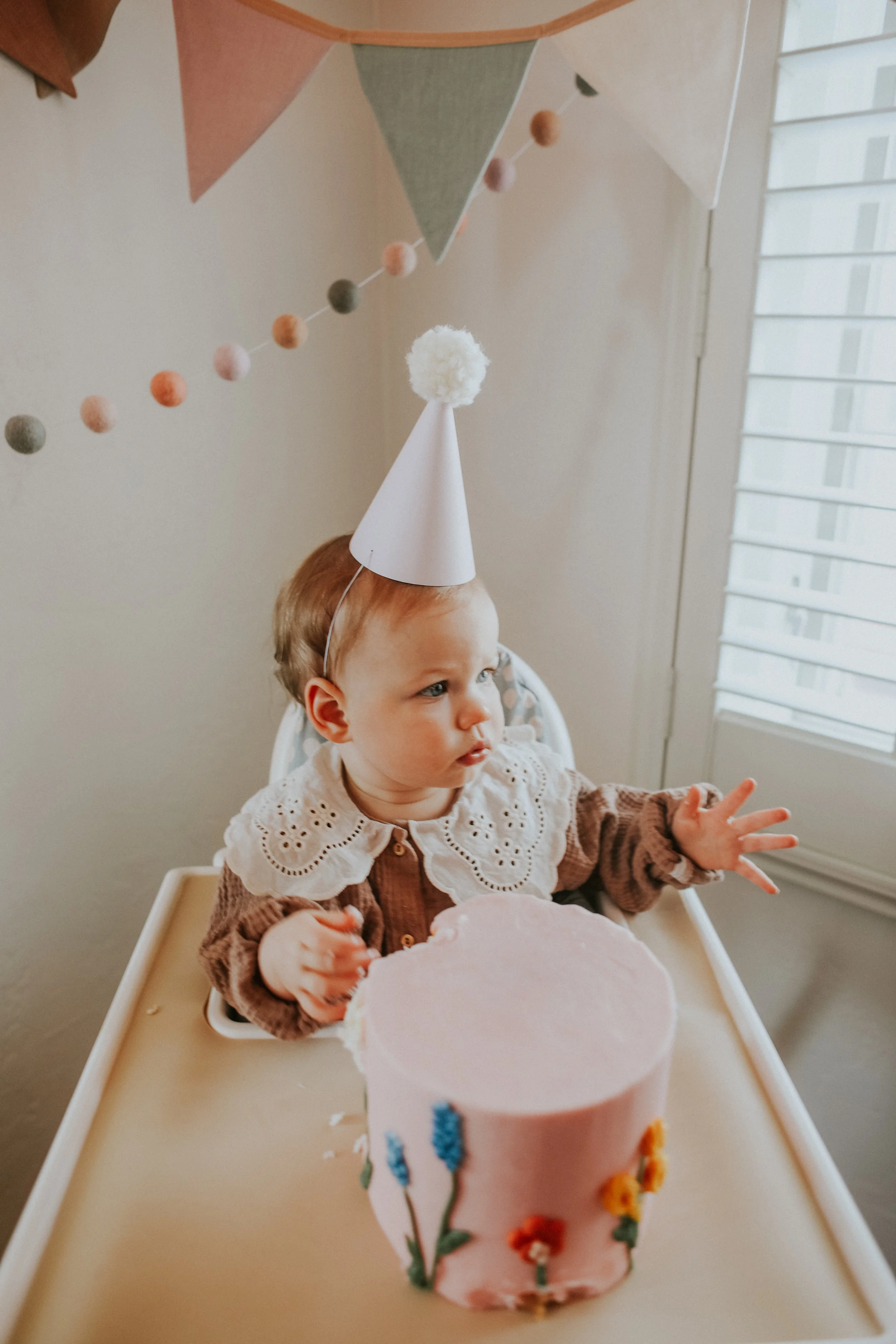 A young child wearing a white party hat with a pom-pom on top, sitting at a high chair with a pink birthday cake decorated with flowers, celebrating a birthday.