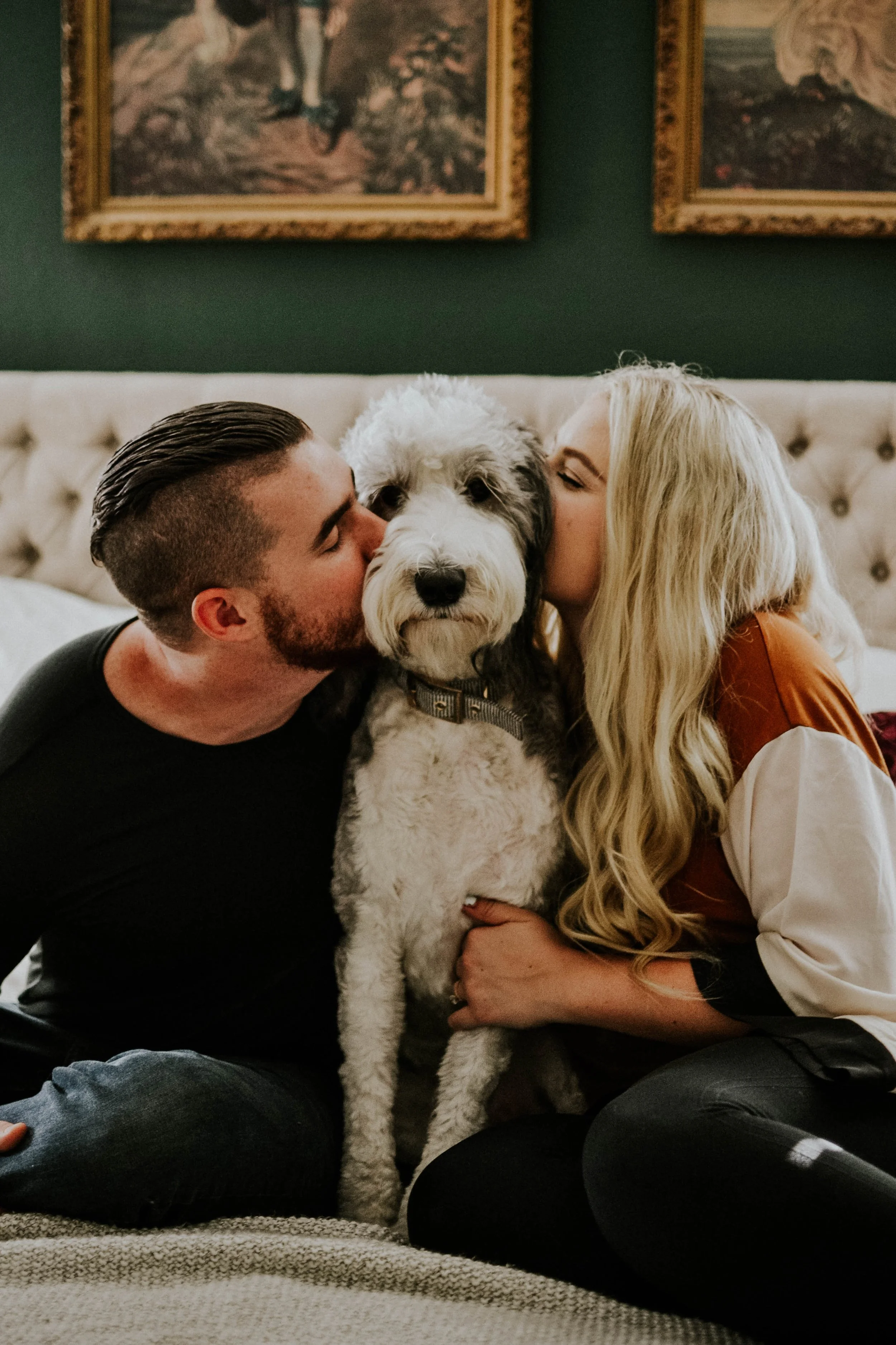 A couple is kissing their dog on the cheeks while sitting on a bed in a cozy, decorated room.