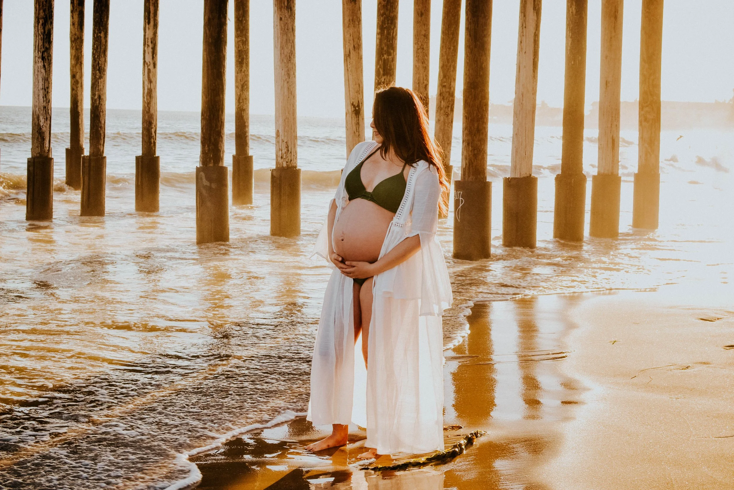 A pregnant woman standing barefoot on a sandy beach near wooden pier pillars, wearing a bikini top and light, open cover-up at sunset.