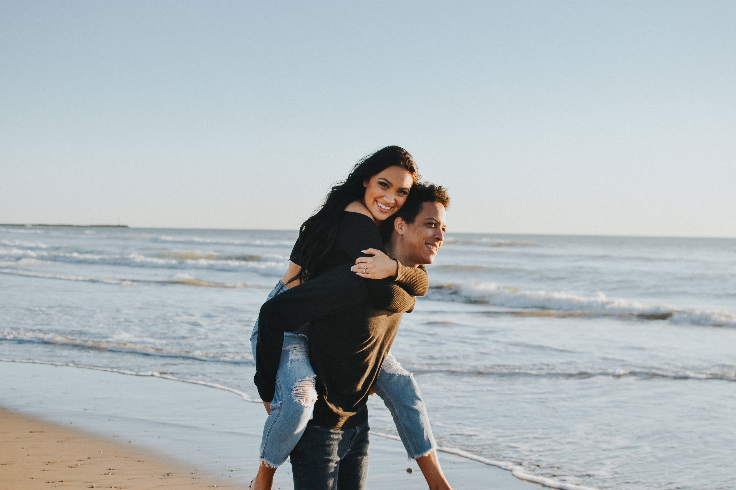 A couple enjoying the beach, with the woman sitting on the man's back as they smile and look at each other, on a sandy shore with ocean waves in the background.