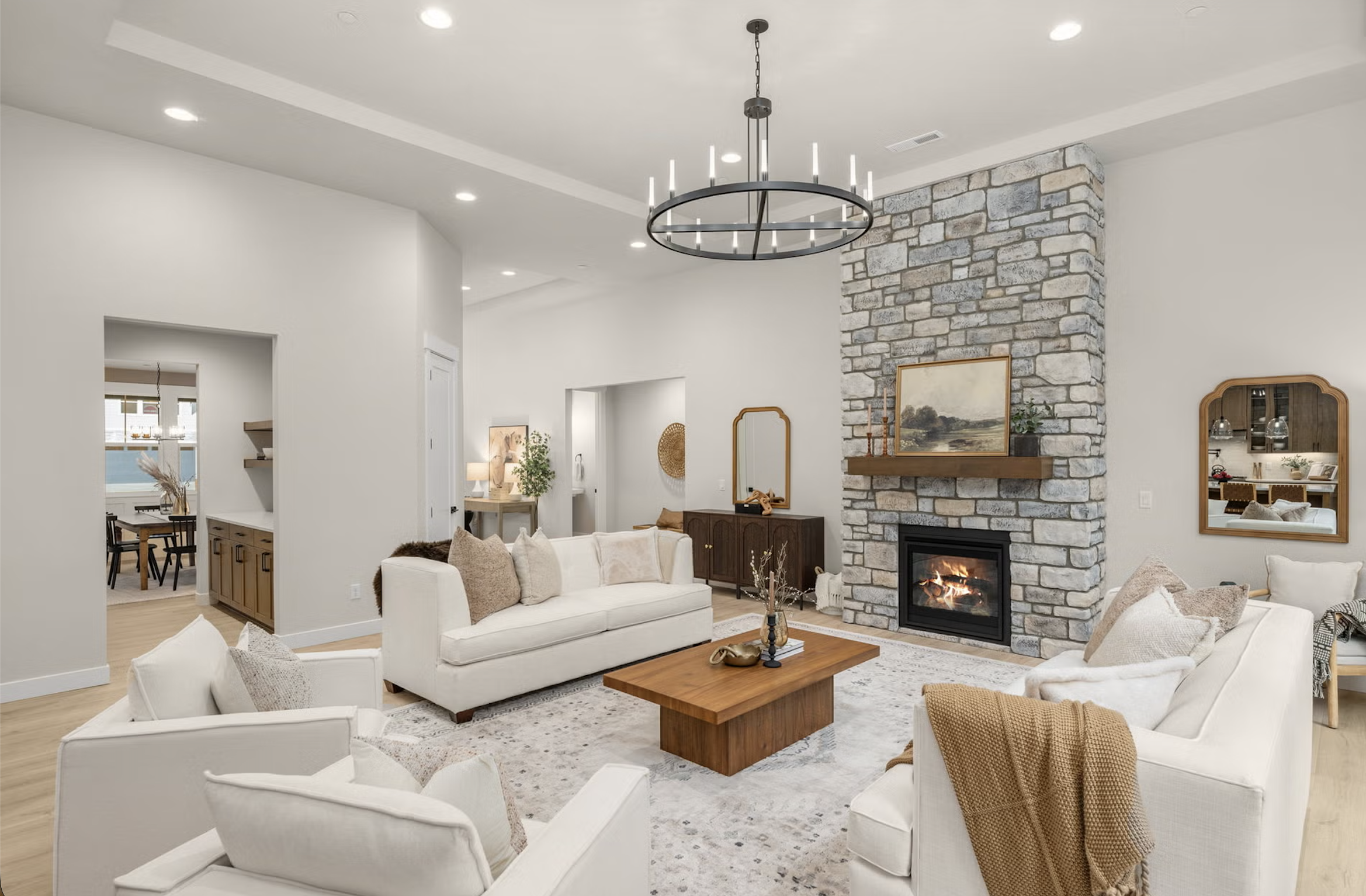 Living room with white sofas, a wooden coffee table, and a fireplace with a stone chimney. Decor includes artwork, mirrors, and lamp, with an open view to the dining area.
