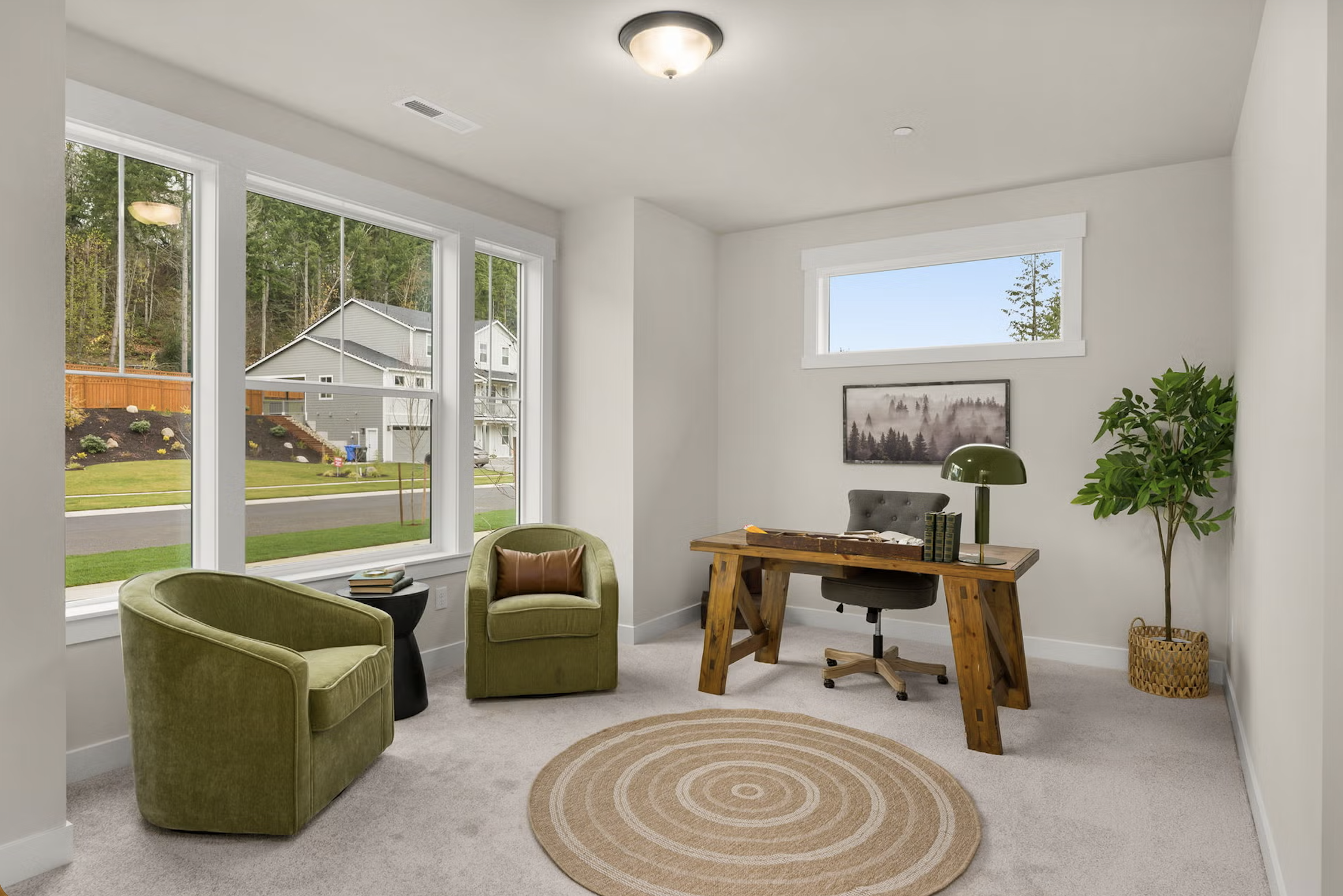 A modern home office with large windows, a wooden desk, a black office chair, a green armchair and a matching green side chair, a small round beige rug with circular designs, a potted plant, and a framed landscape picture on the wall.