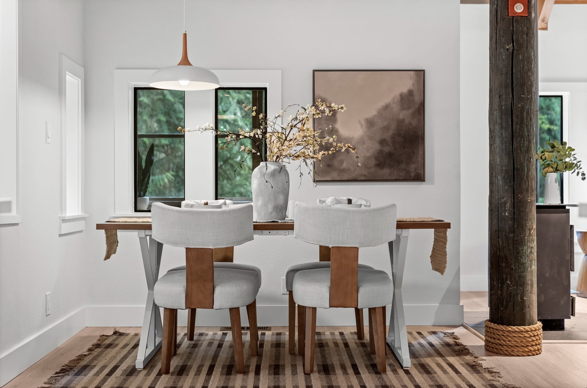 Dining room with a wooden table, two white and wood chairs, a large gray vase with white flowers, a painting, and a pendant light. Windows in the background reveal greenery outside.