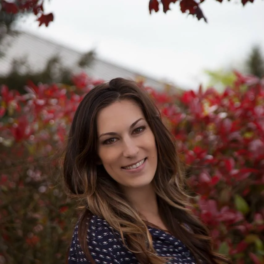 A woman with long, wavy brown hair smiling outdoors, standing in front of a bush with red leaves.