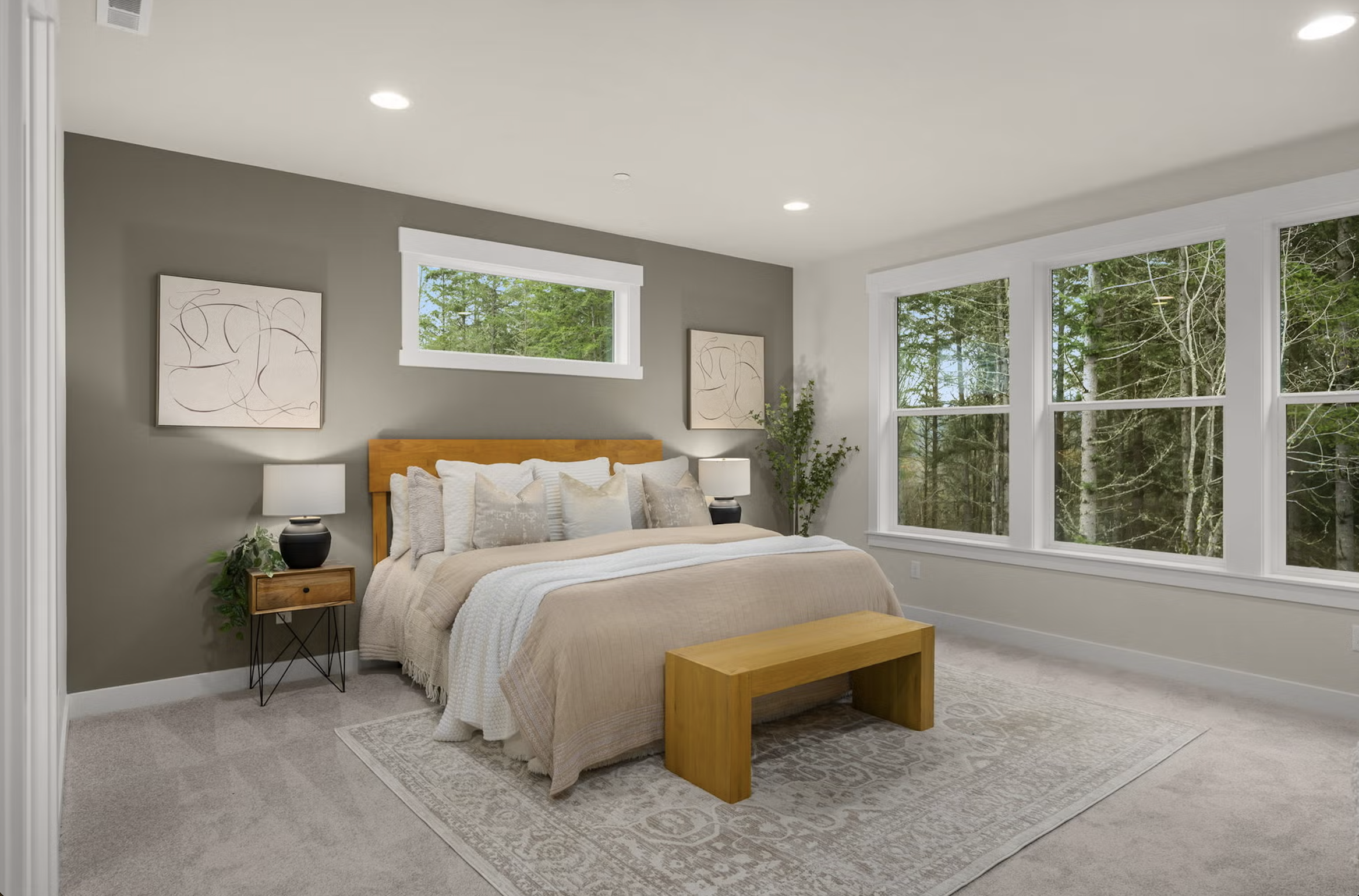 A modern bedroom with a bed, wooden headboard, and matching bench, beige bedding, two nightstands with lamps, and large windows showing a wooded outdoor view.