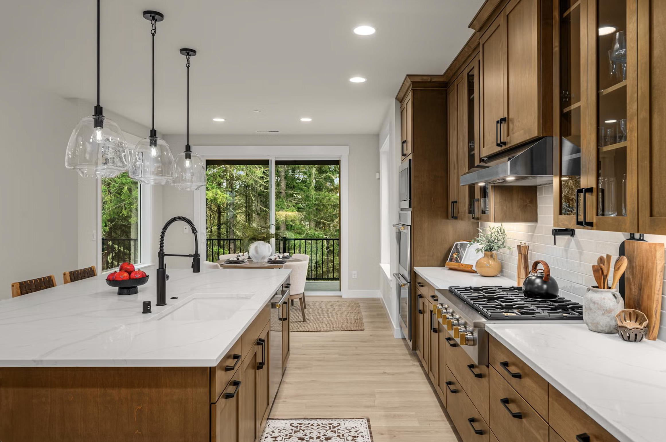 Modern kitchen with wooden cabinets, white marble countertops, and a large island with a black faucet. There are three glass pendant lights above the island, and a sliding door leading to a balcony with trees outside. The kitchen has a stove, oven, microwave, and decorative items such as a black teapot, potted plant, and wooden utensils.