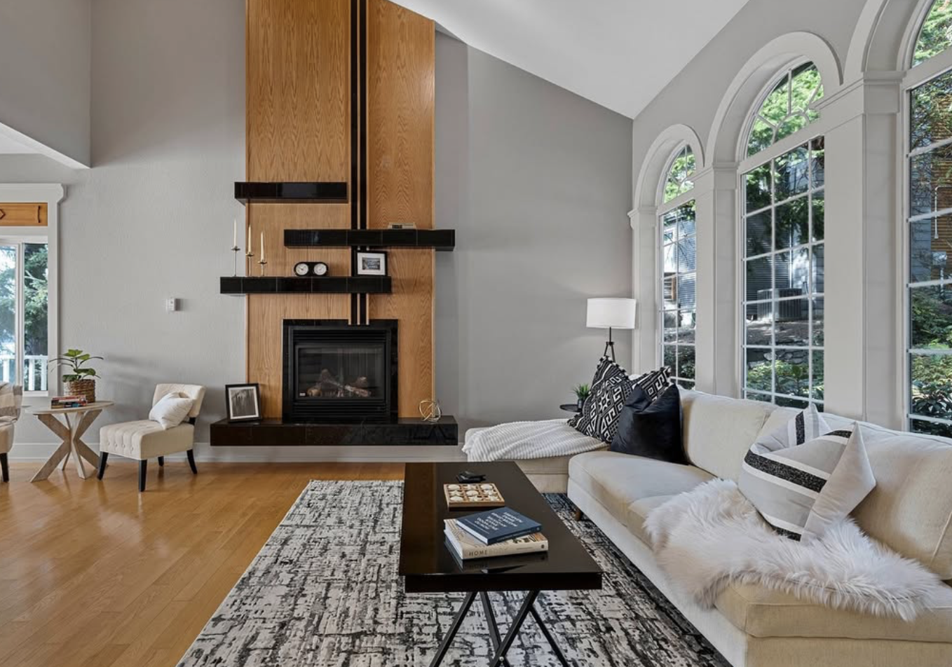 Living room with large arched windows, white sofa with black and white pillows, fireplace with wooden and black accents, coffee table with books, and hardwood floors.