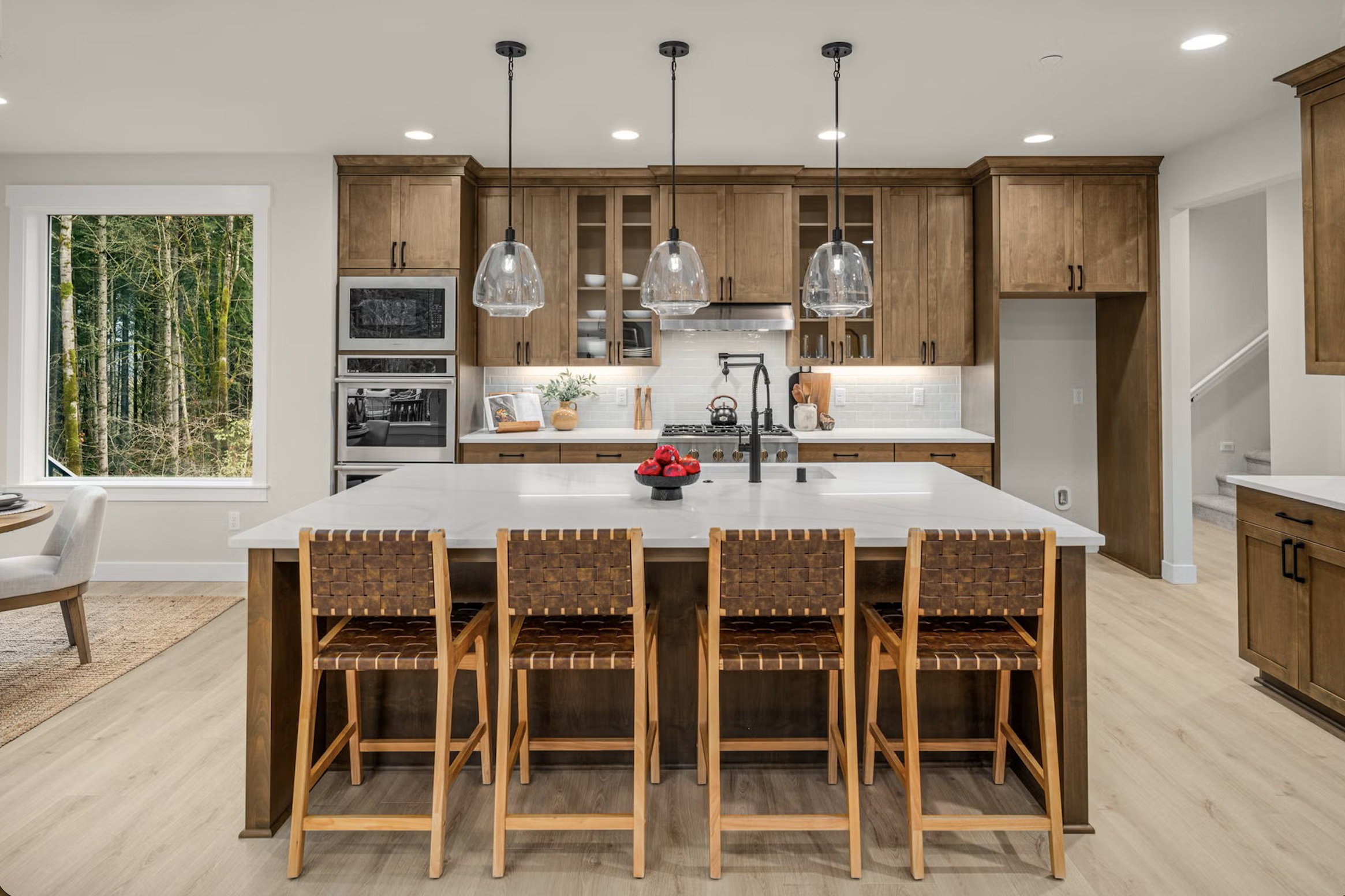 Modern kitchen with wooden cabinets, white countertops, and a large island with five woven-seat chairs. Hanging glass pendant lights over the island, and a window showing a forest scene outside.