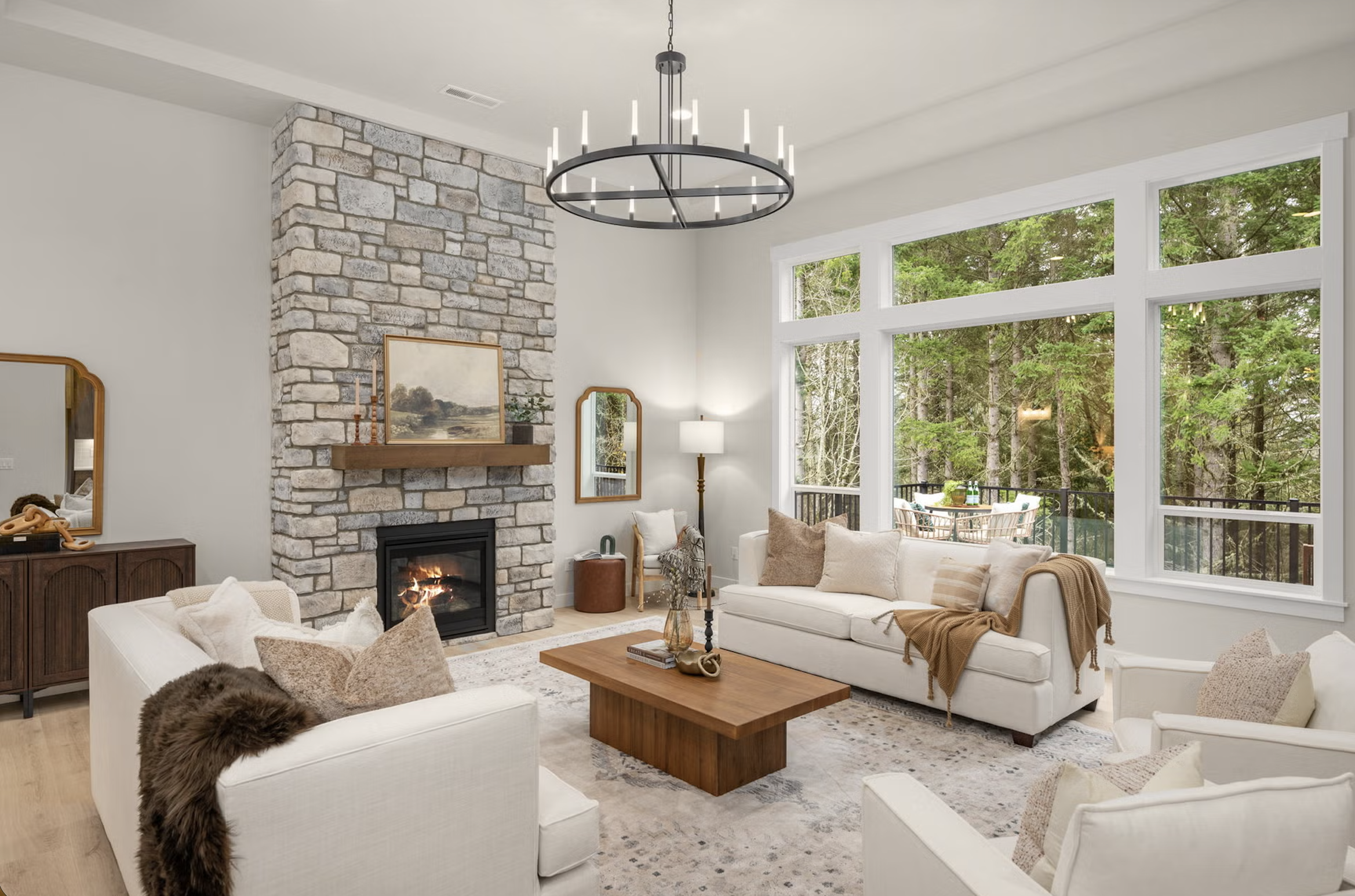 Living room with stone fireplace, white sofas, large windows showing trees, and wooden coffee table.