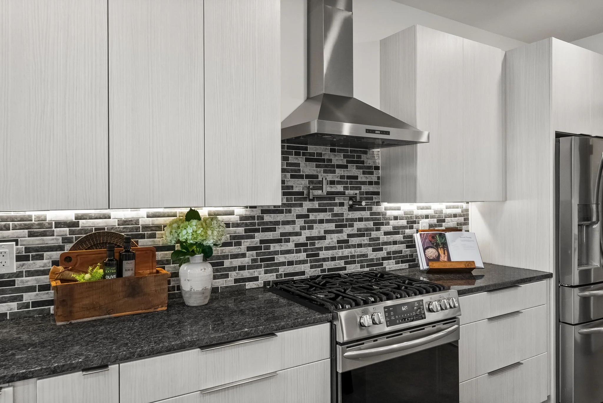 Modern kitchen with white cabinets, a black granite countertop, a stainless steel stove with oven, a stainless steel range hood, and a gray tiled backsplash. Decorative items include a wooden tray with soap and lotion bottles, a vase with green and w