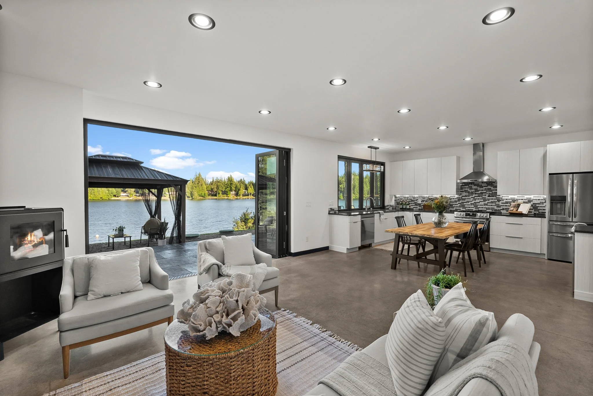 Open-concept living room and kitchen with large windows overlooking a lake, with outdoor seating, modern furniture, fireplace, and kitchen appliances.