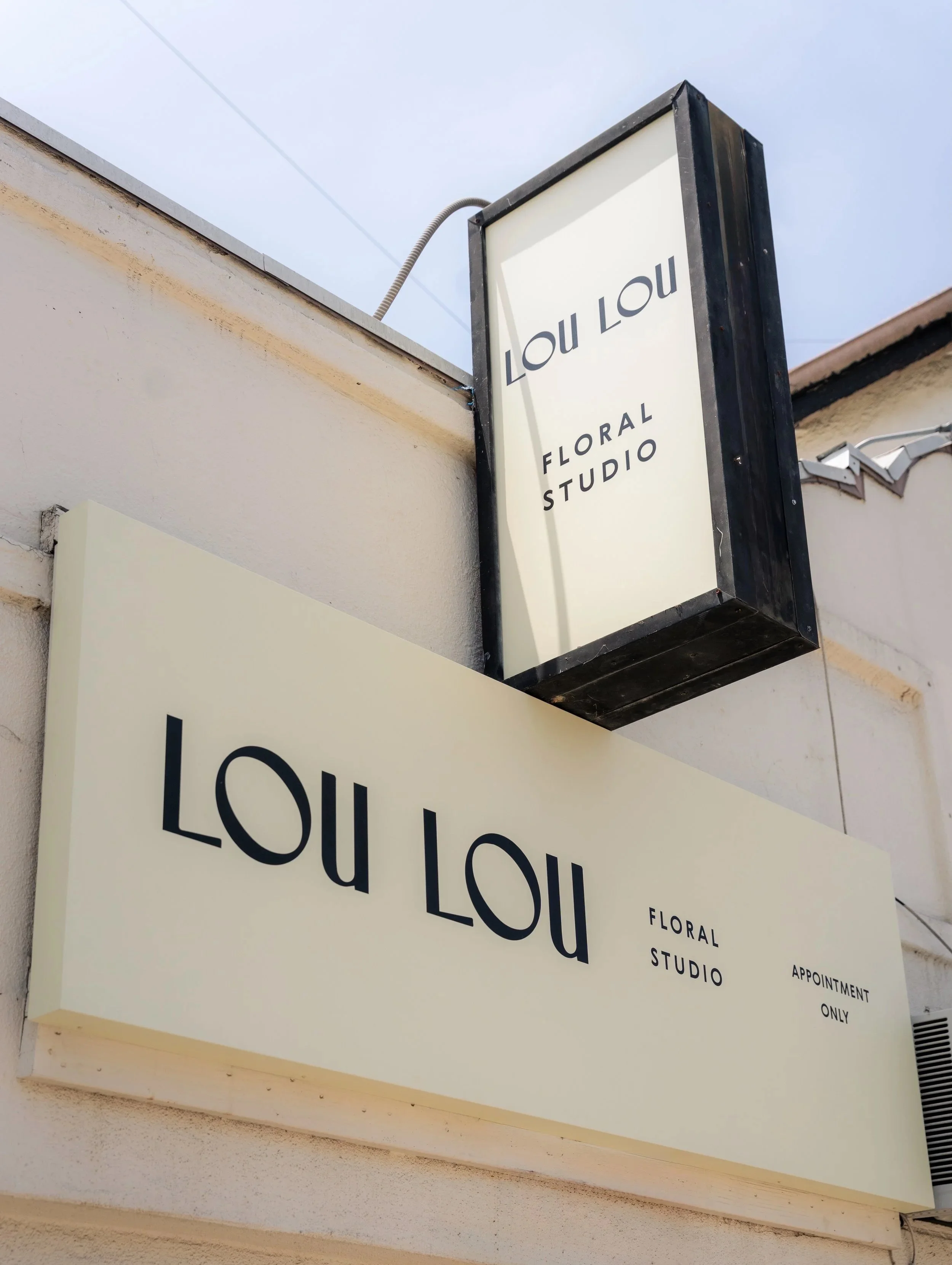Sign for Lou Lou Floral Studio with black border, white background, and black text, mounted on building exterior.