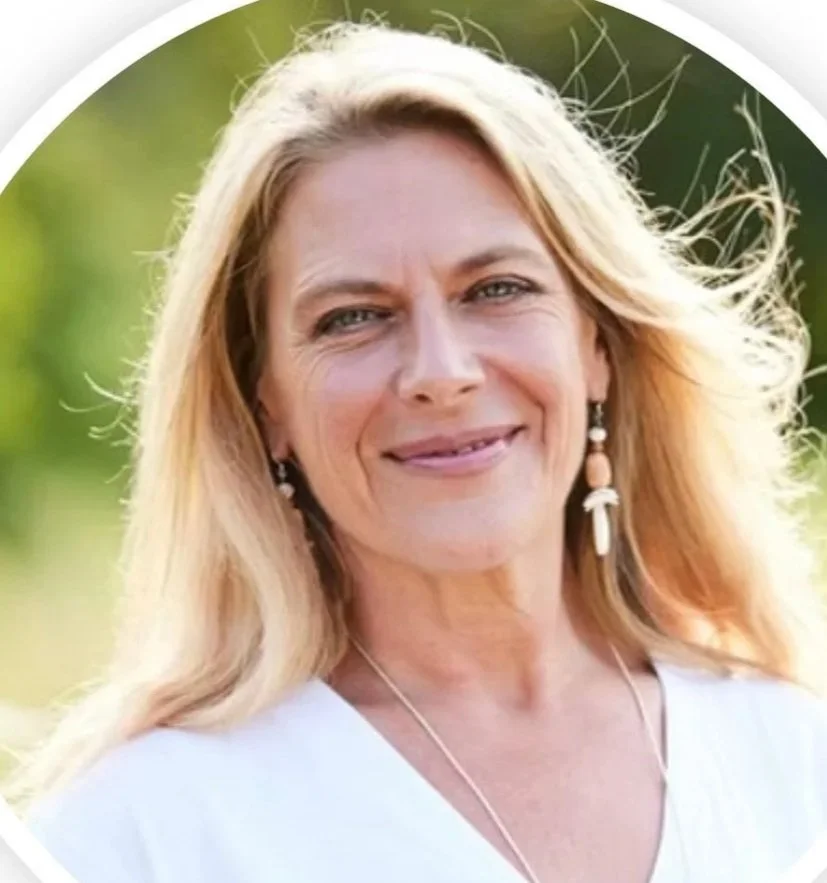 A middle-aged woman with blonde hair, wearing earrings and a white top, smiling outdoors with blurred greenery in the background.