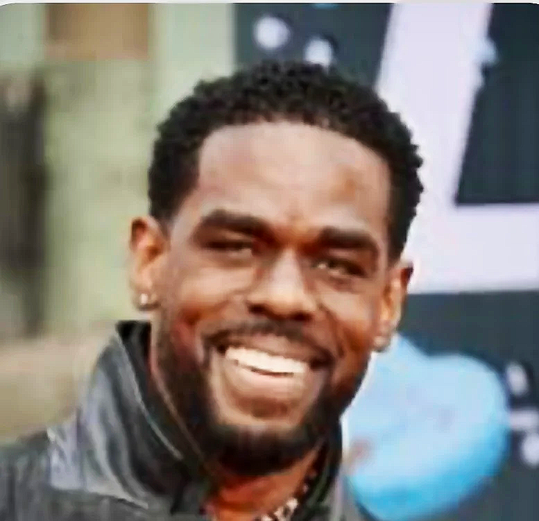 Close-up of a smiling man with curly hair, wearing earrings and a leather jacket.
