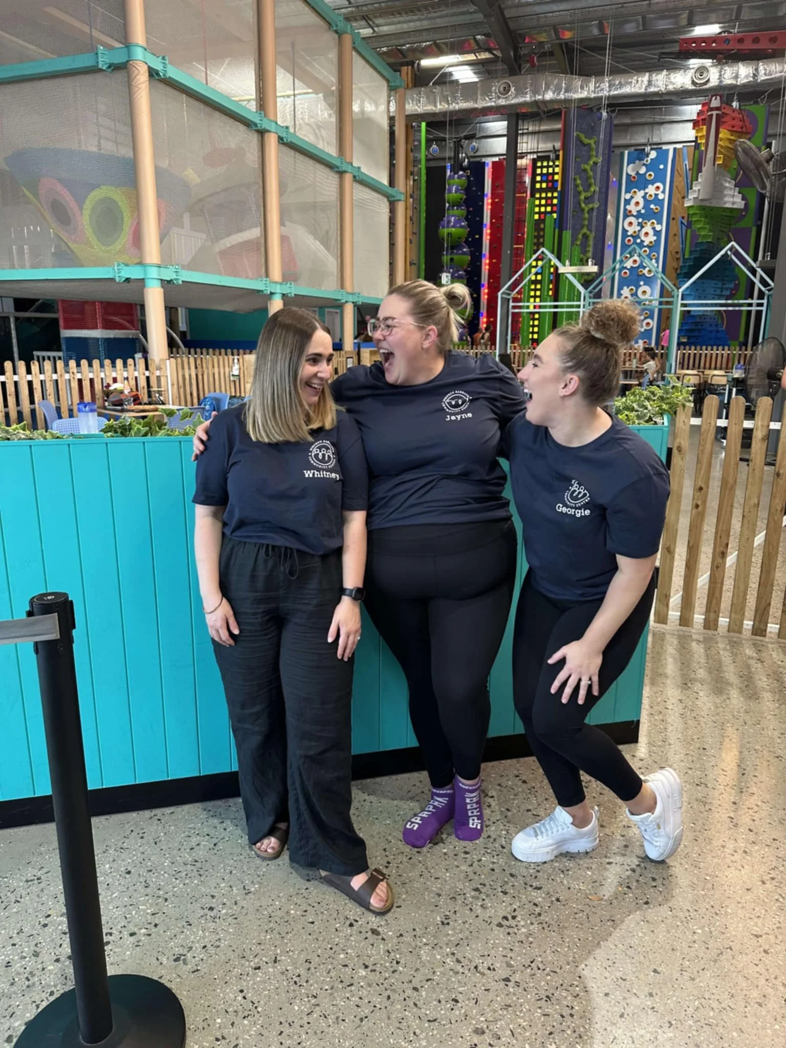 Three women wearing matching navy shirts with names and logos, standing together inside an indoor adventure park, smiling and laughing with arms around each other, behind a teal-colored fence and colorful climbing structures.