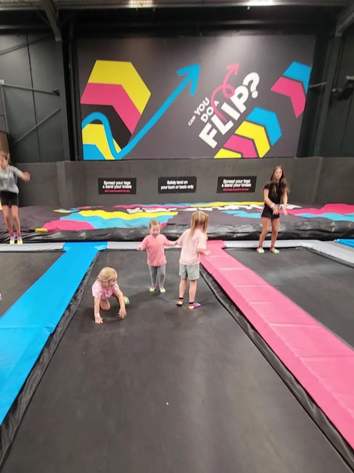 Children playing on trampolines in an indoor trampoline park with colorful wall art and bouncing mats.