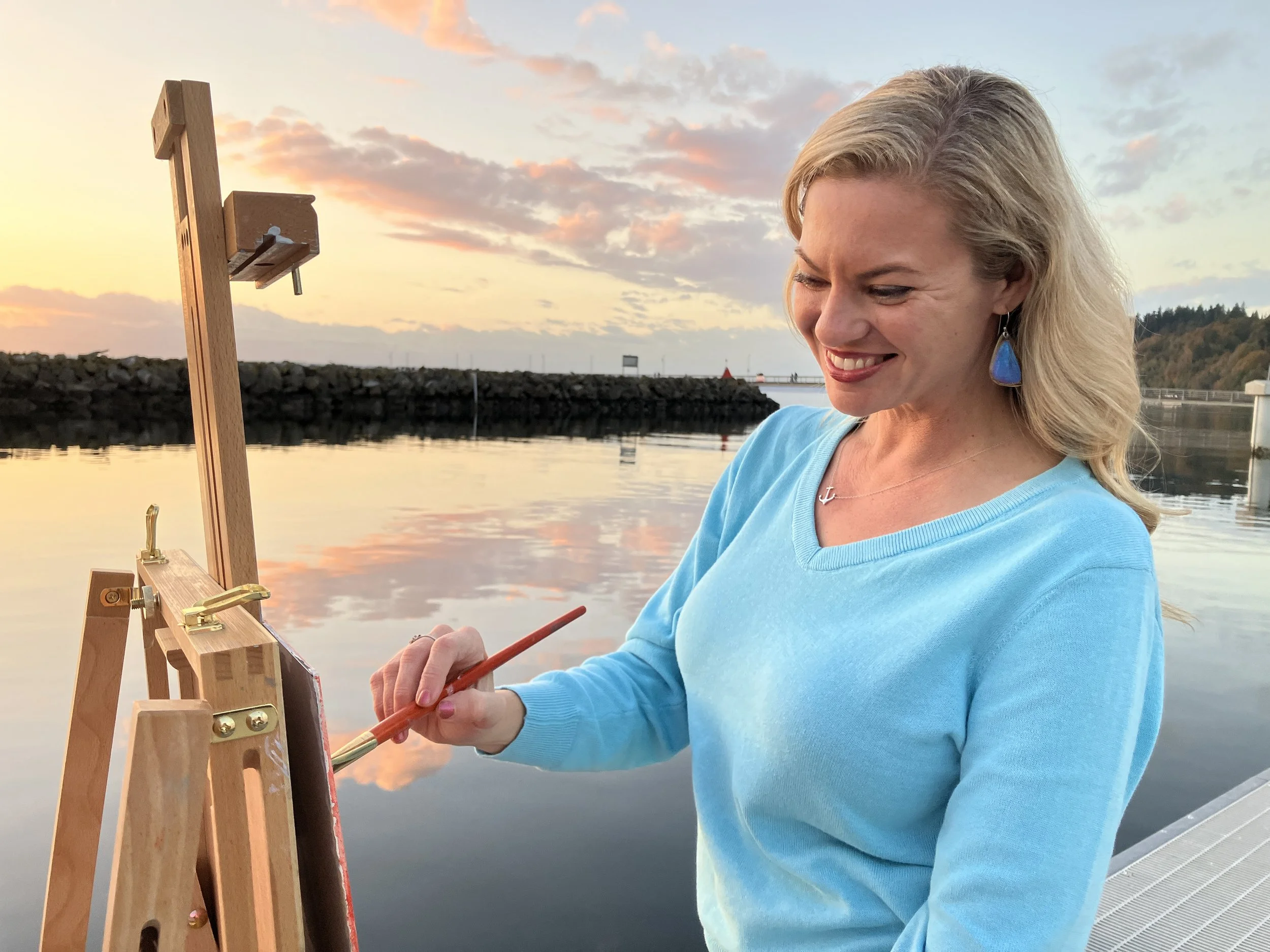 Woman painting outdoors near water during sunset, wearing a blue sweater and blue earrings, smiling as she holds a paintbrush.