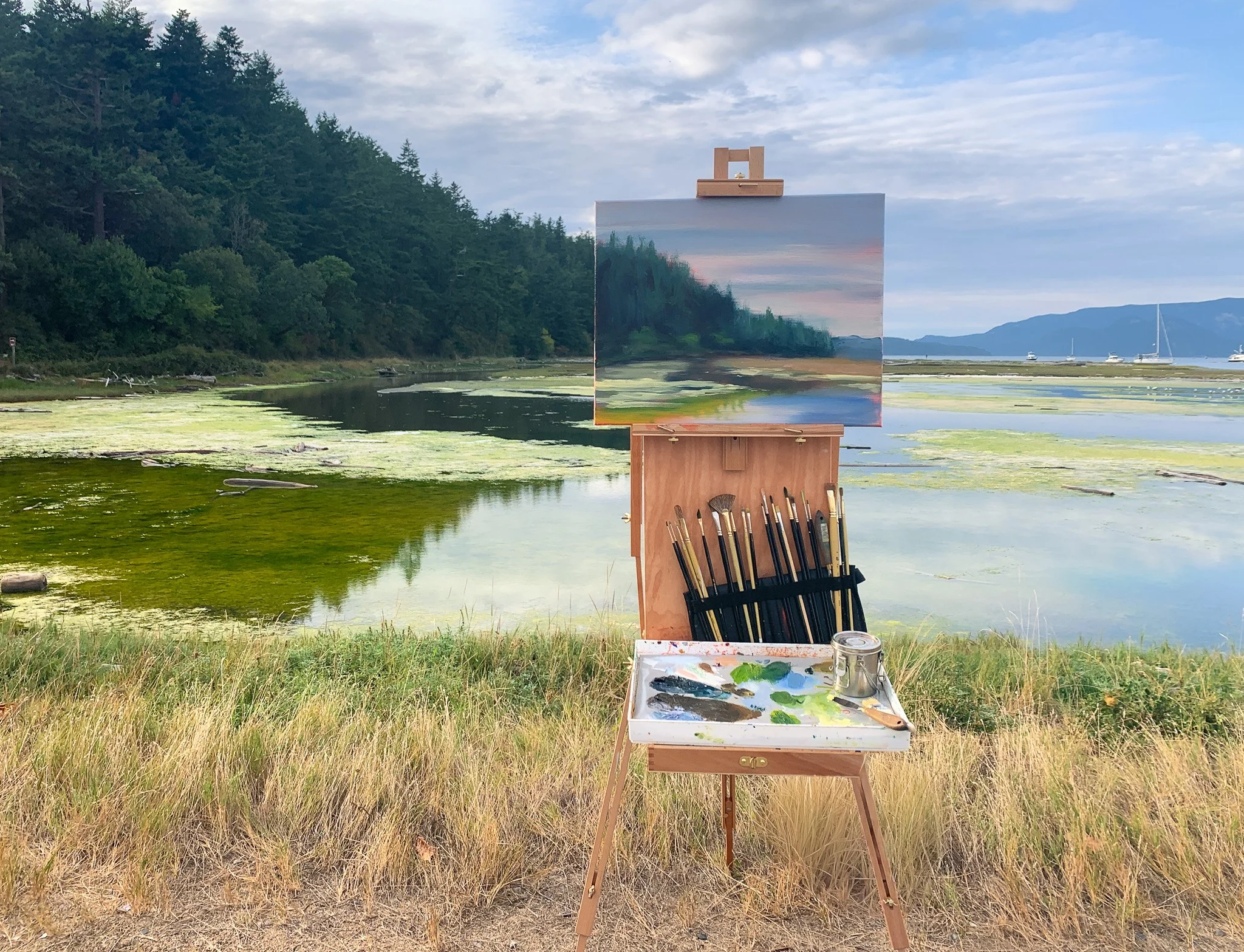Outdoor scene with an easel holding a landscape painting of a lake and trees. Art supplies and brushes are on a tray at the base of the easel, surrounded by grass near a body of water, with a forested hillside in the background and a cloudy sky.