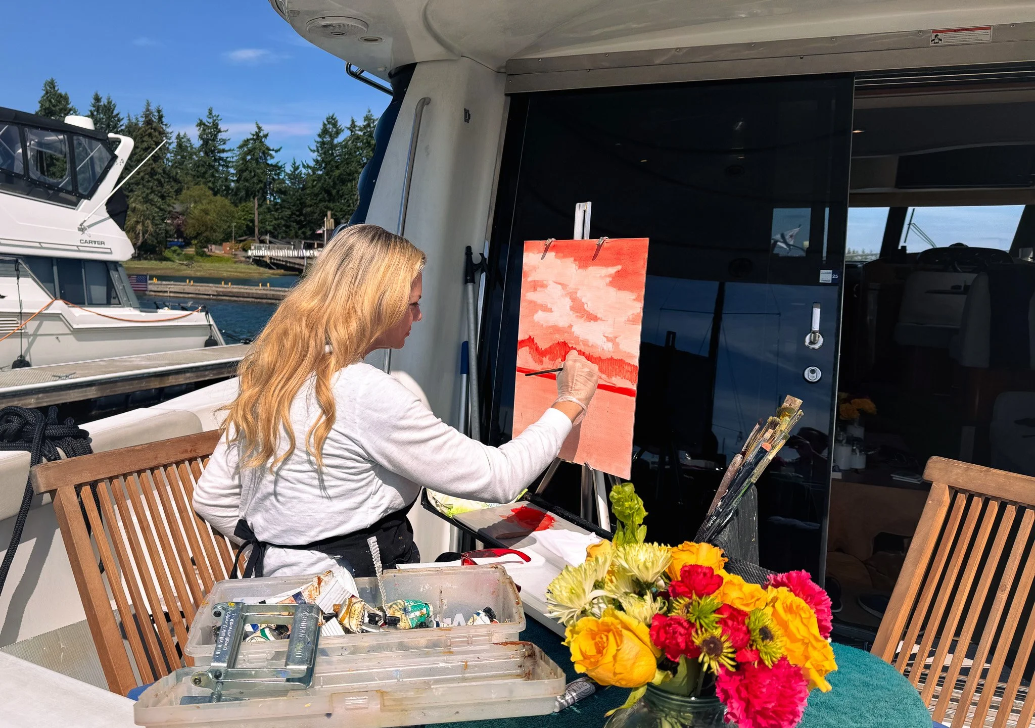 A woman with long blonde hair painting a landscape on a canvas outdoors on a boat docked at a marina, with parked boats and trees in the background, and a bouquet of colorful flowers on the table.