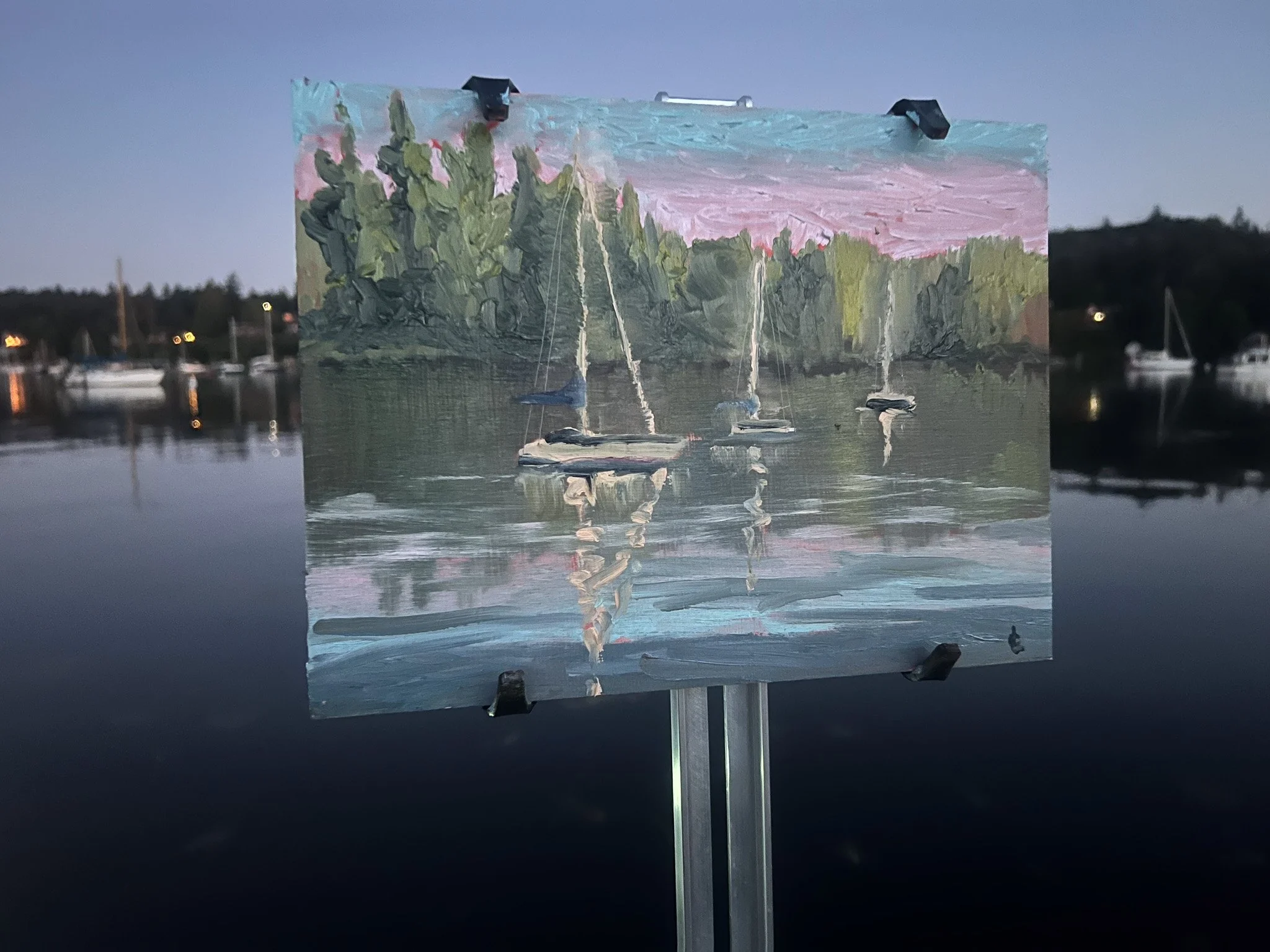 Painting of boats on a calm water lake with trees and a colorful sky in the background, mounted on an easel outdoors near a marina at dusk.