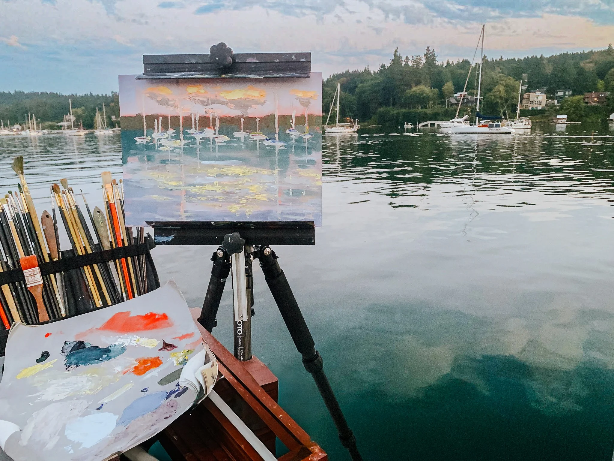 An artist's painting of sailboats on the water on an easel, set up on a dock by a calm lake with sailboats and a wooded shoreline in the background.
