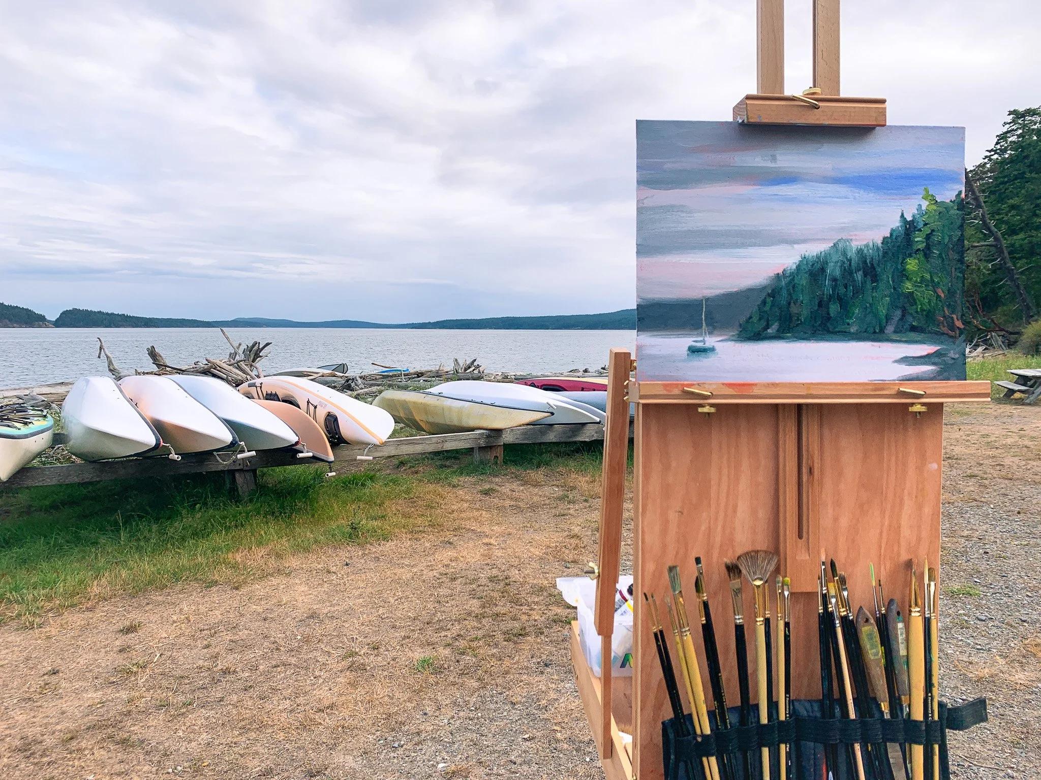 Outdoor scene with a wooden easel holding a landscape painting of a boat on a lake with a forested shoreline, surrounded by kayak boats on the shore and a large body of water under a cloudy sky.