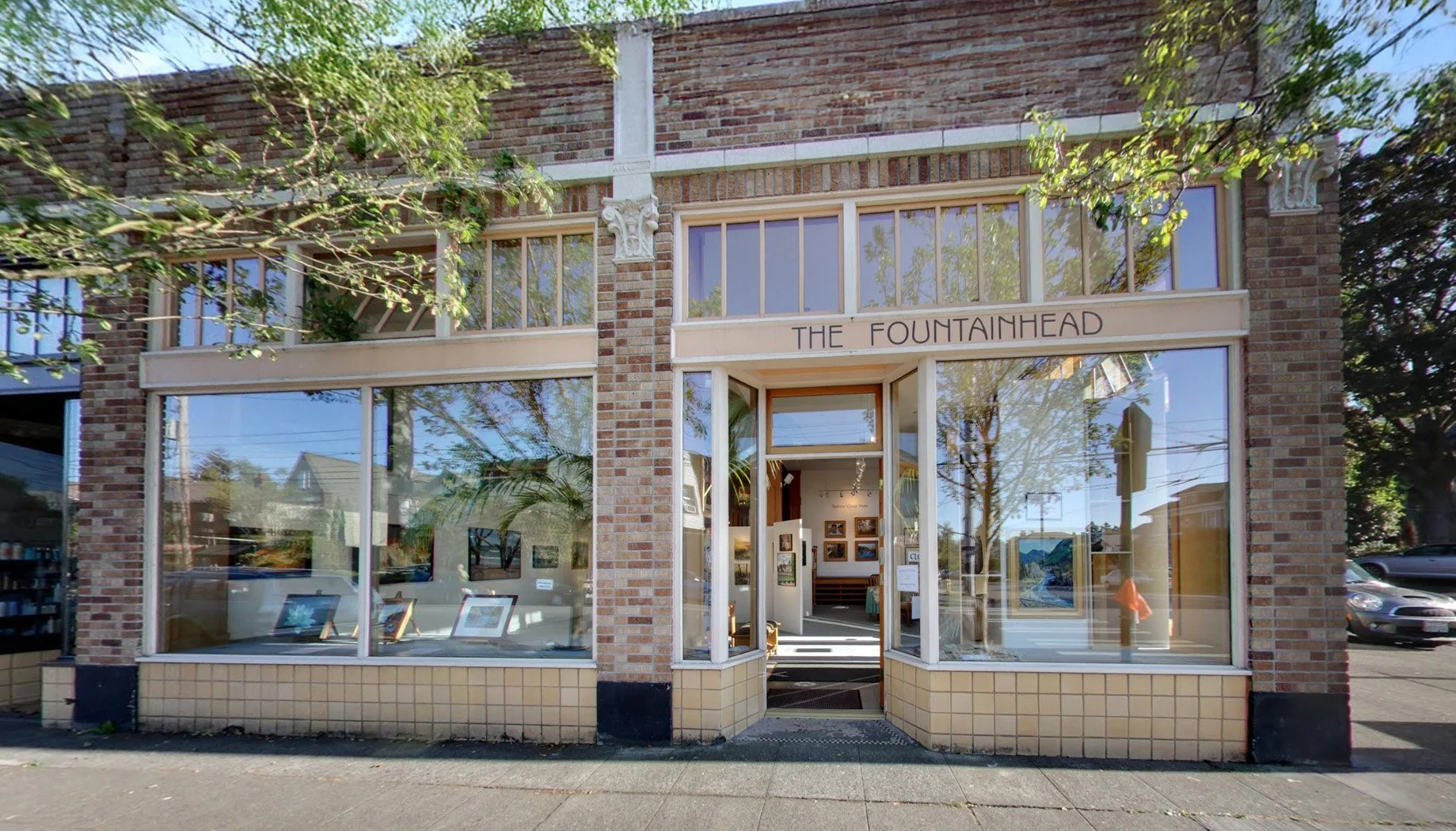 Storefront with large windows displaying framed artwork, above the entrance it says "The Fountainhead" in capital letters, brick exterior with decorative white trim.