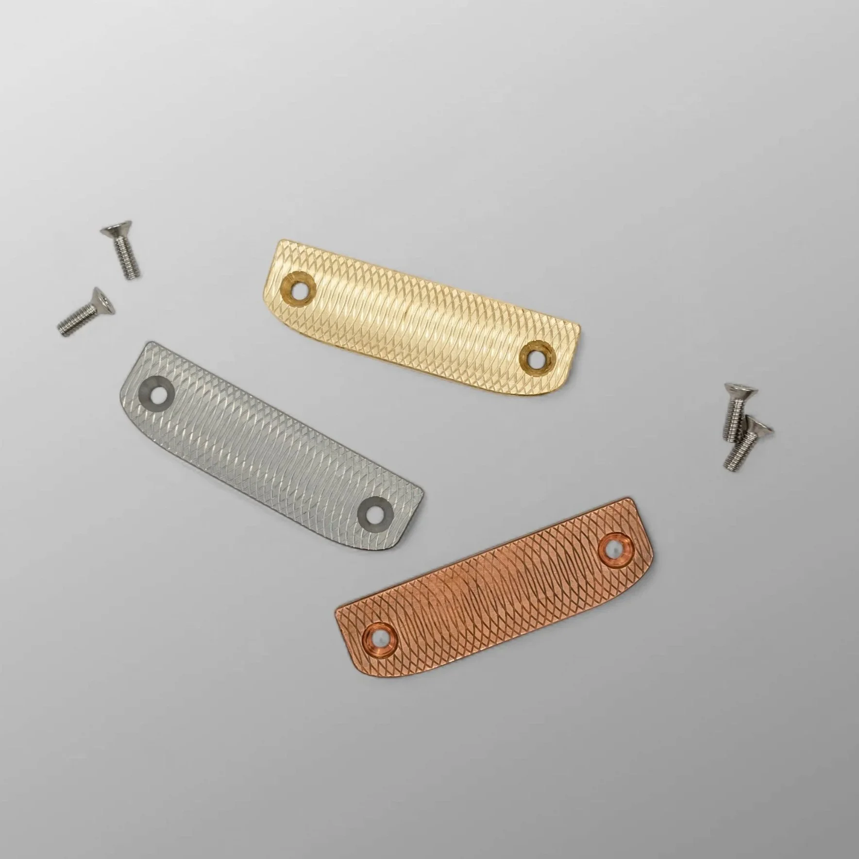 Three metallic putter face plates in brass, stainless steel, and copper with textured surfaces and holes, along with two screws on a light gray background.