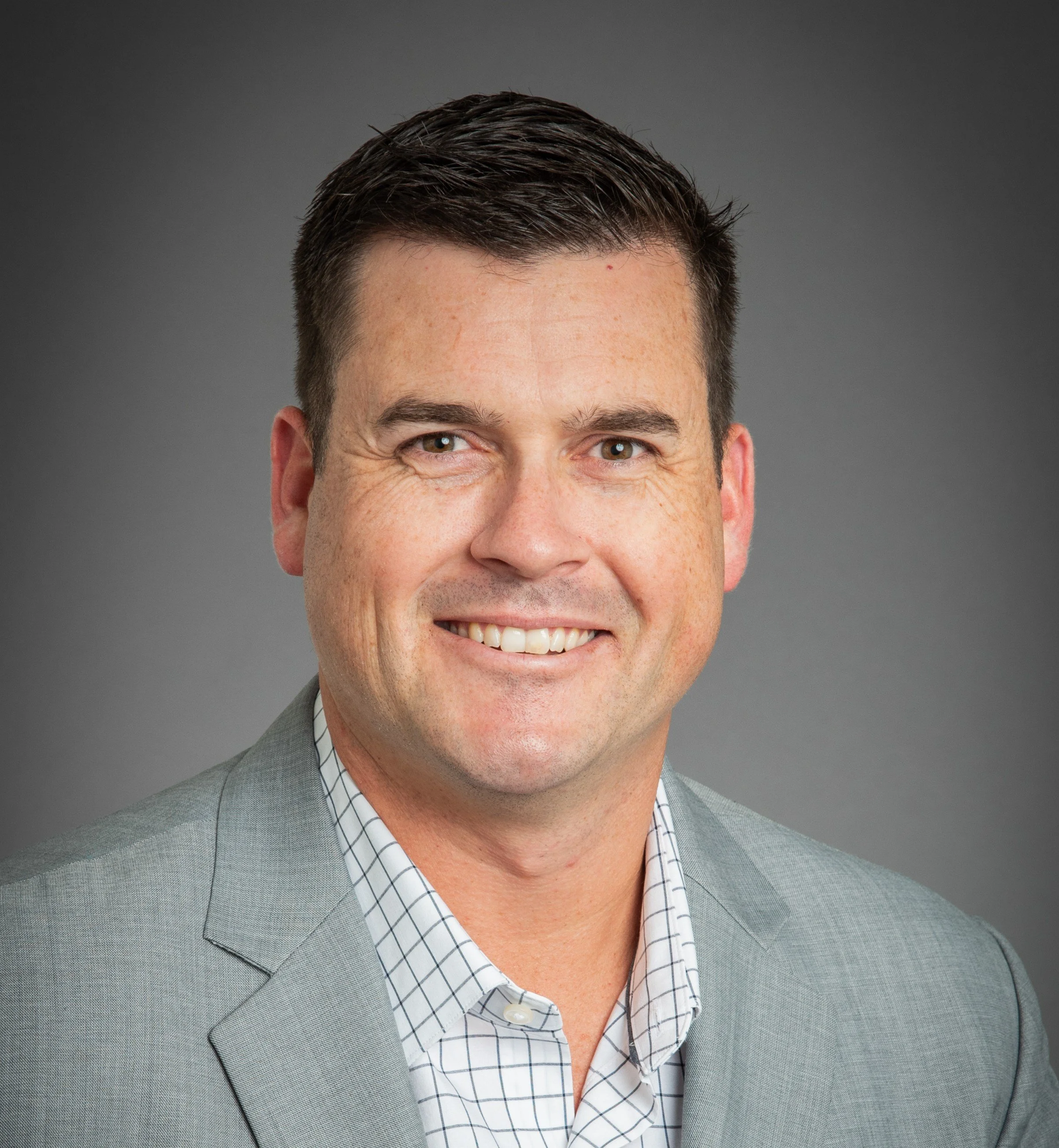 Professional headshot of Blaine Worthington, Founder and Executive Director, a smiling man with short dark hair, wearing a light gray suit and a checkered dress shirt, against a gray background.