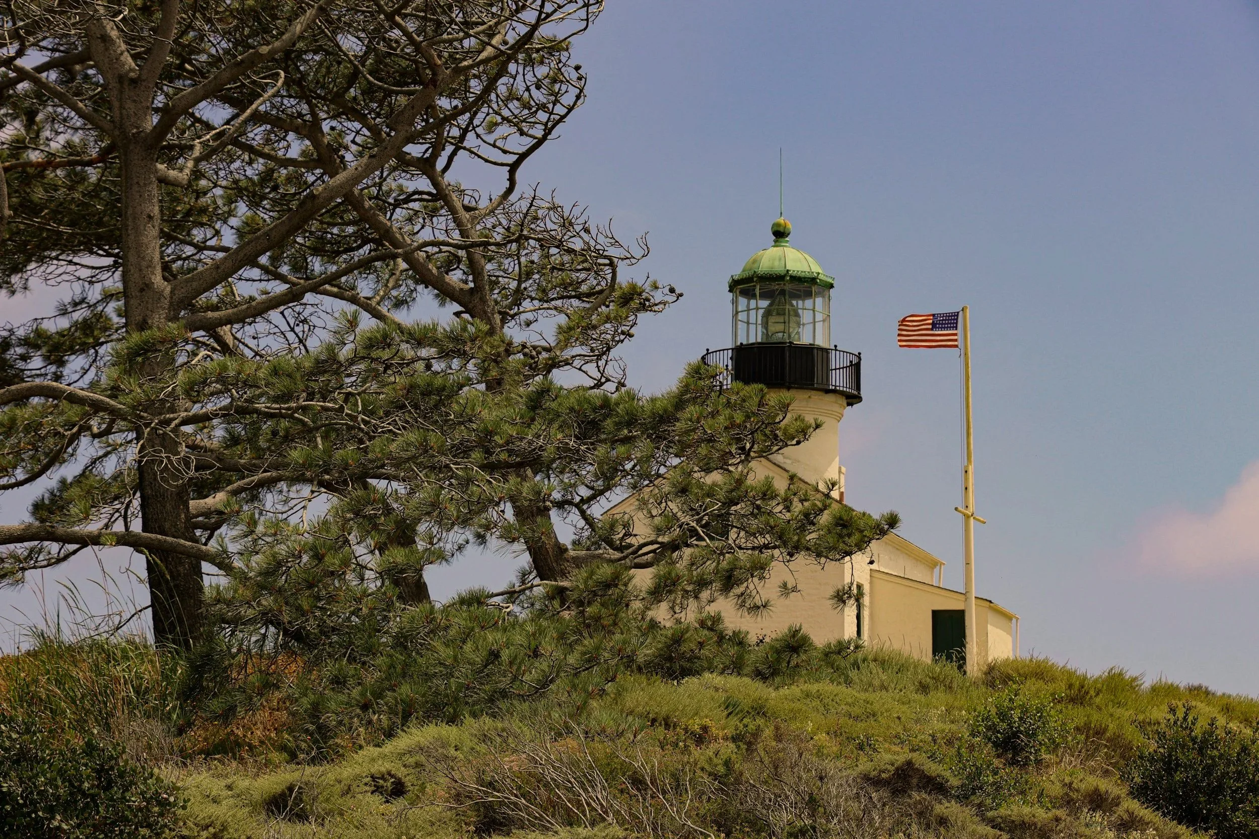A lighthouse with a green dome, situated on a grassy hill, with an American flag flying on a flagpole nearby. There are trees in the foreground and a blue sky with a few clouds in the background.