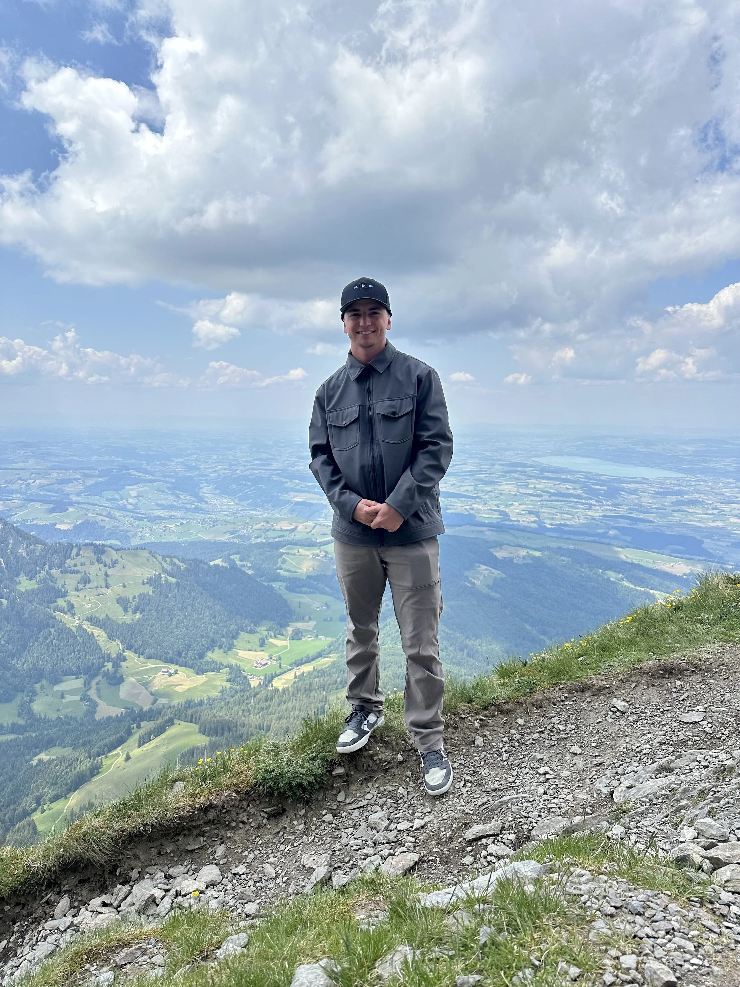 A young man standing on a mountain trail with a scenic view of valleys, forests, and lakes under a partly cloudy sky.