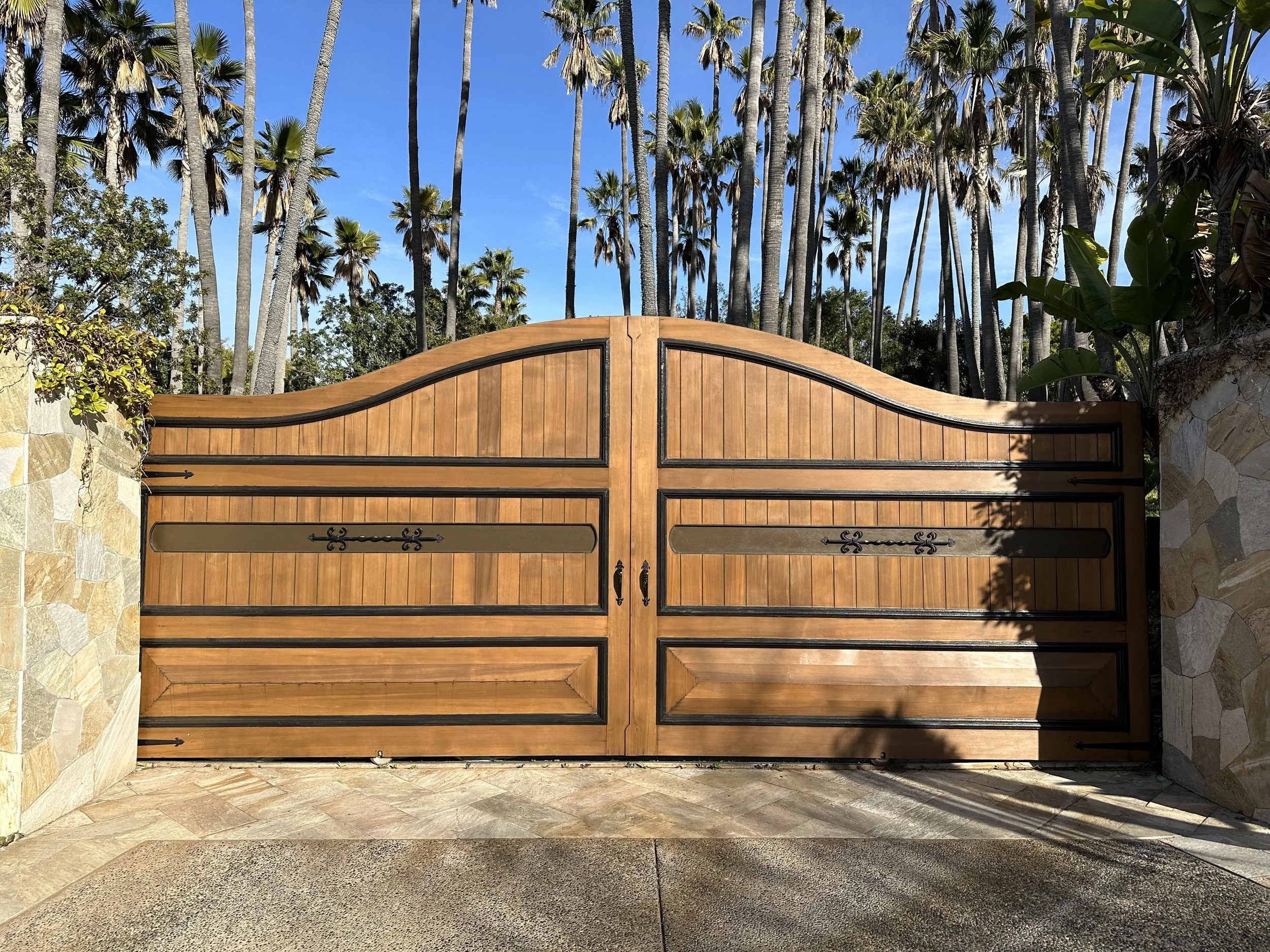 A large wooden gate with black decorative iron hardware, set between stone pillars, with tall palm trees and a blue sky in the background.