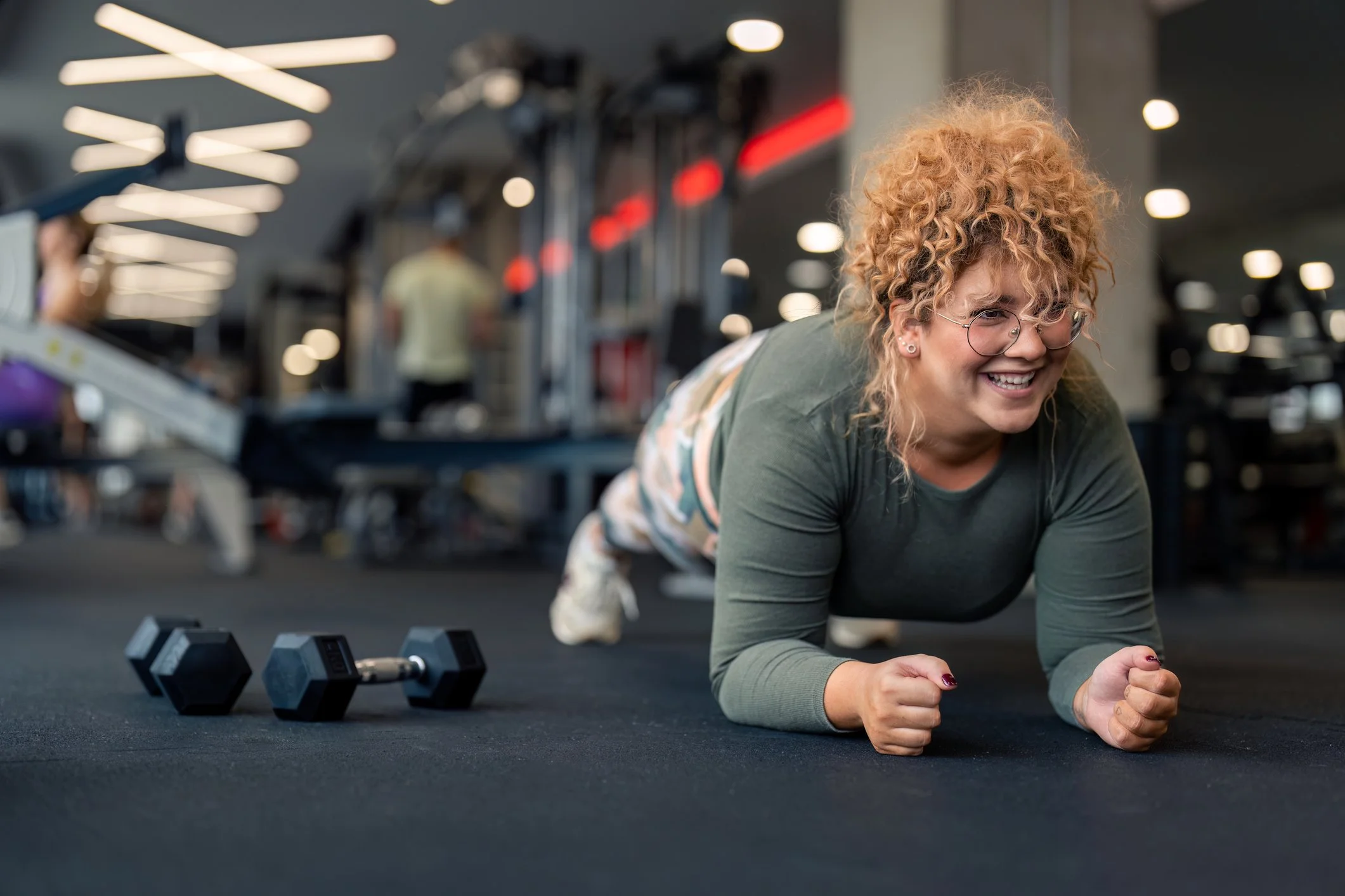 A woman with curly red hair and glasses doing a plank exercise on the gym floor, with a smile on her face, and a pair of dumbbells nearby.
