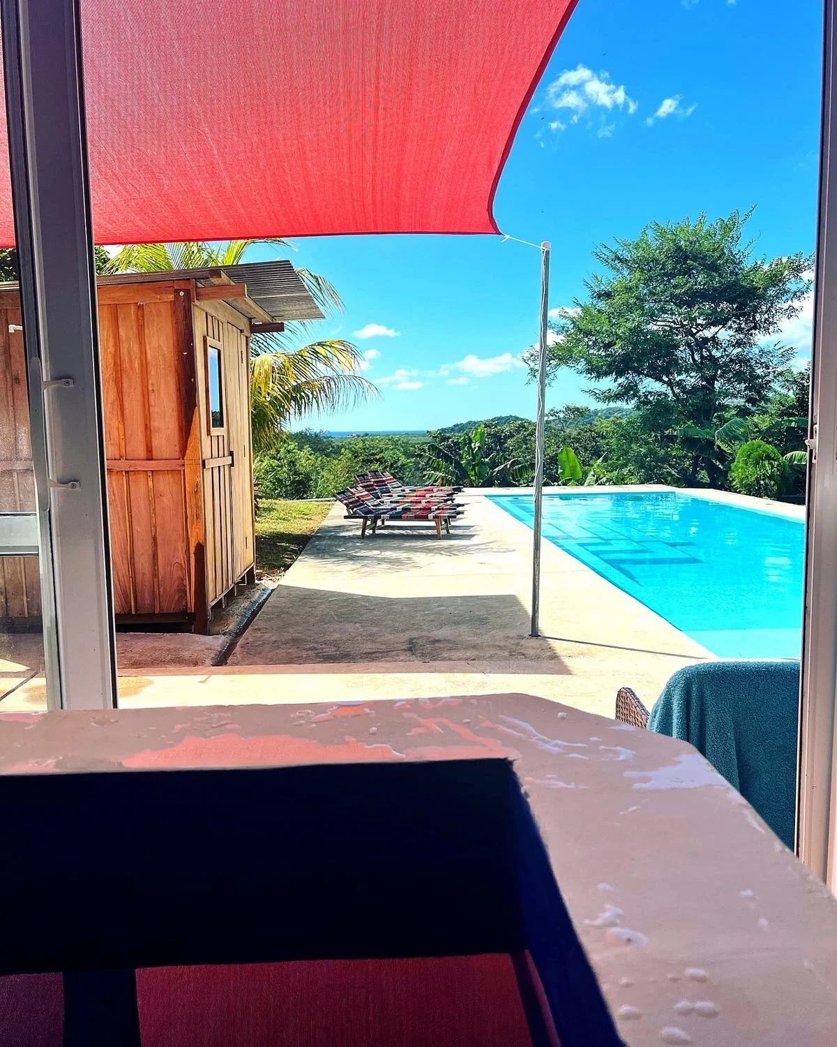 View from near a window opening to an outdoor pool area with lounge chairs, green trees, and a bright blue sky with clouds.