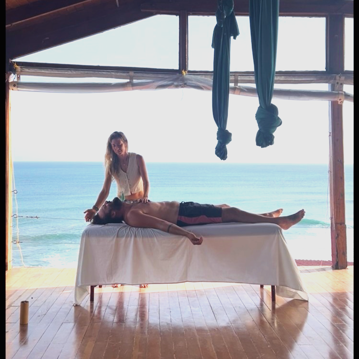 A woman giving an energy session to a man lying face-up on a massage table with an ocean view in the background.