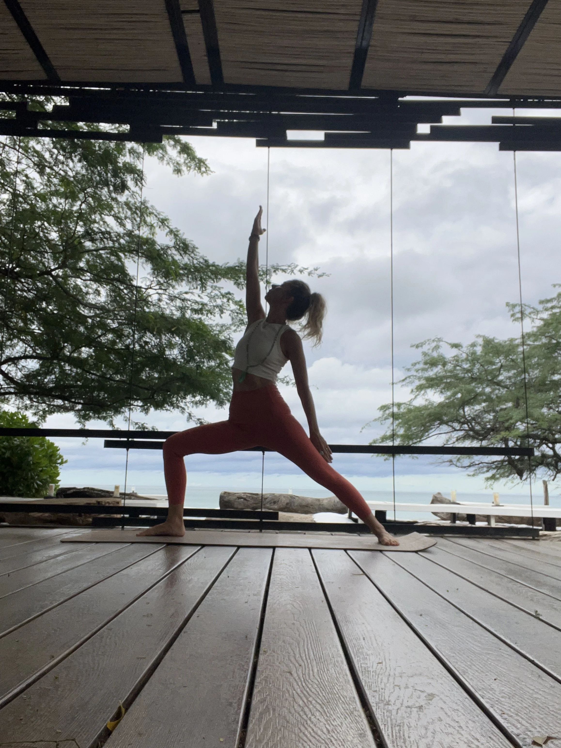 A woman practicing yoga on a wooden deck by the beach, performing a warrior pose with one arm extended upward and the other hand on her bent knee, facing toward the ocean and cloudy sky.