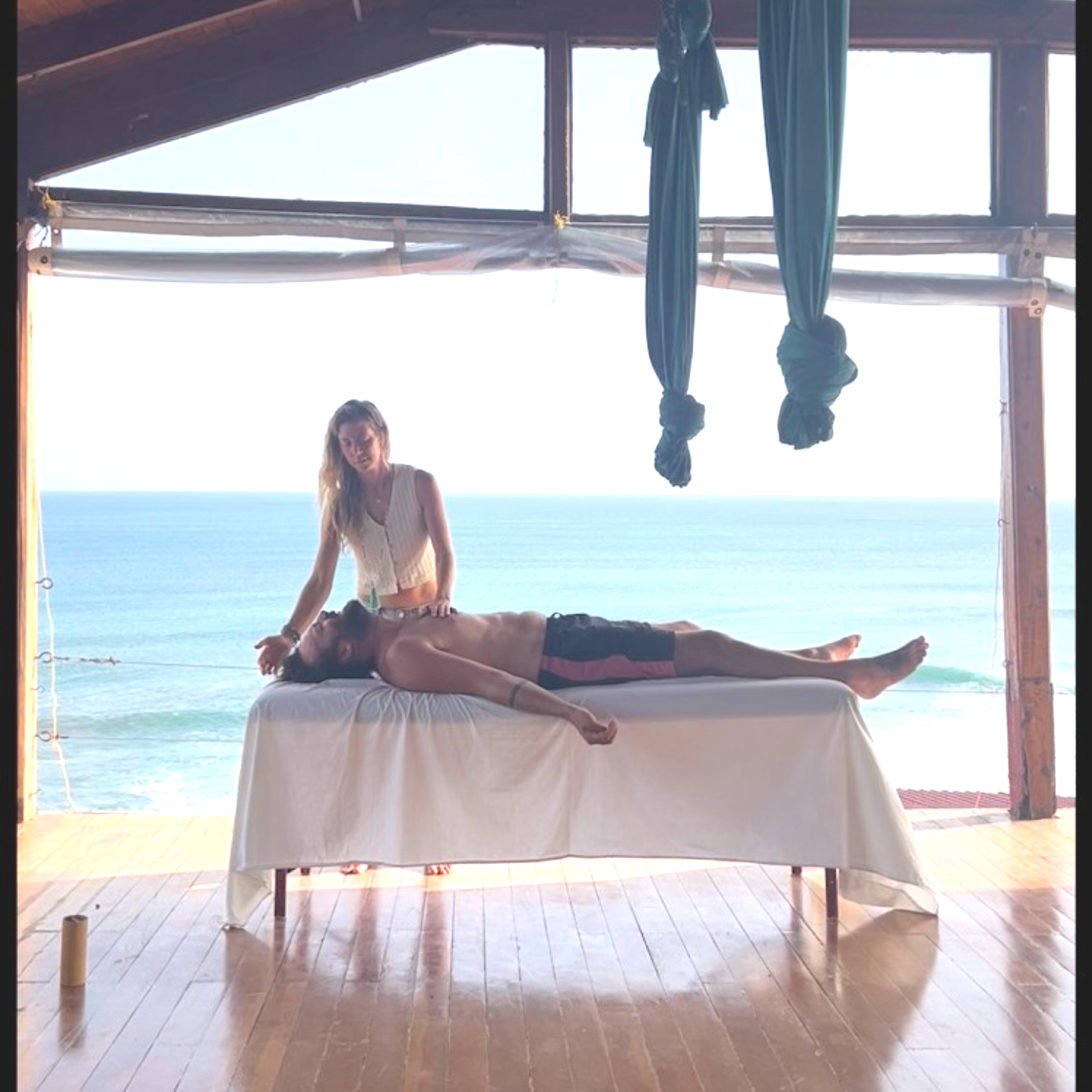 A man receives an energy session on a table with an ocean view in the background, inside a wooden structure.