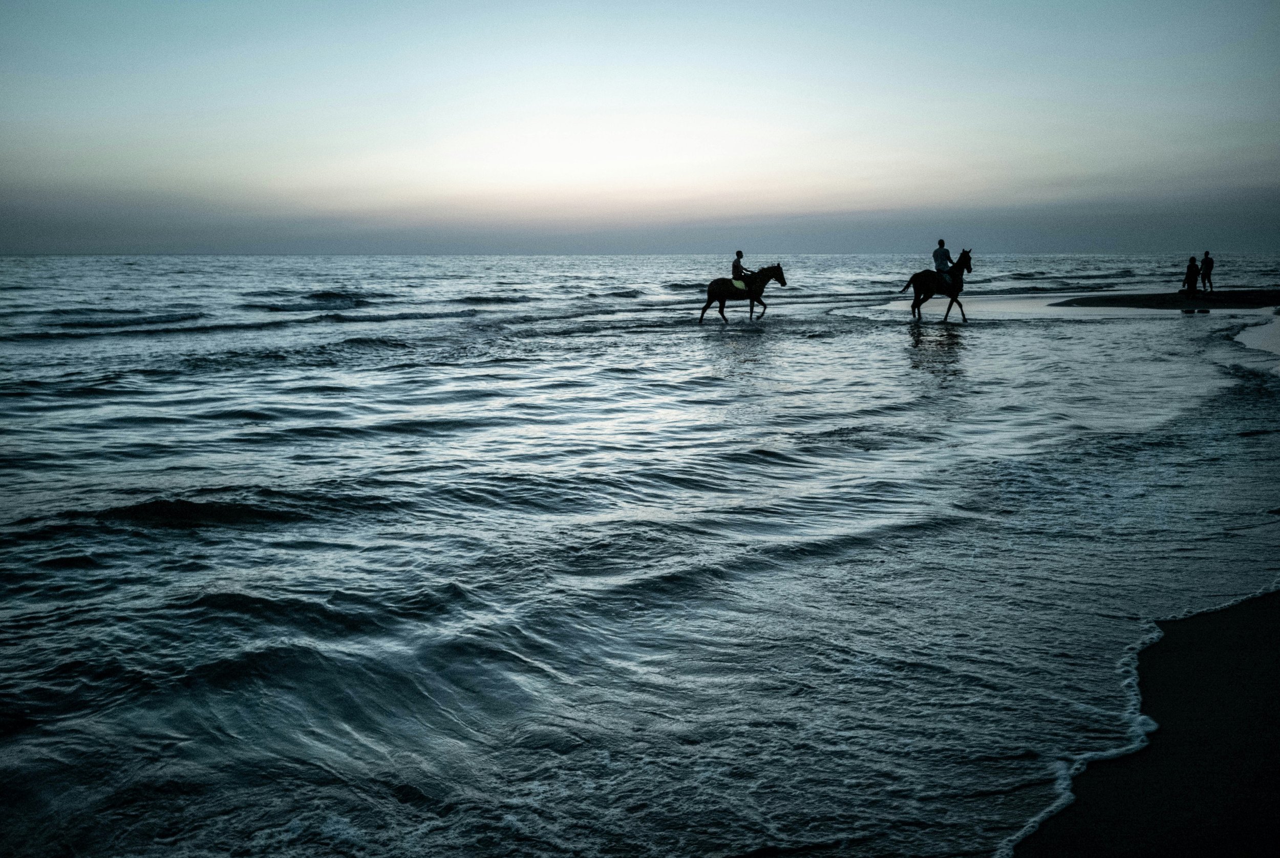 Silhouettes of two people riding horses in the shallow ocean water at dusk or dawn, with two pedestrians walking on the beach in the background.