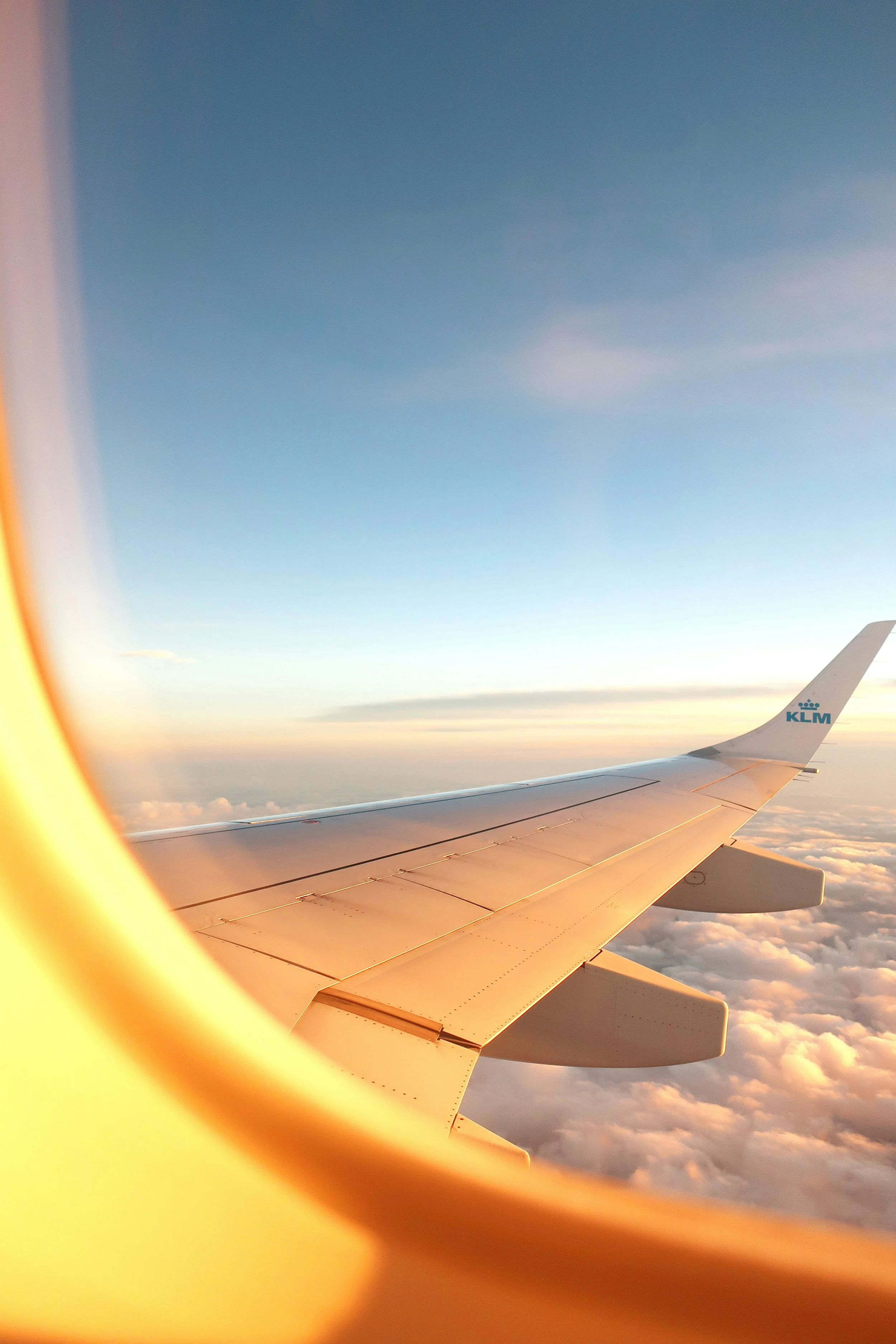 View of an airplane wing seen from a window with the sky and clouds during sunset, with the KLM logo on the wingtip.