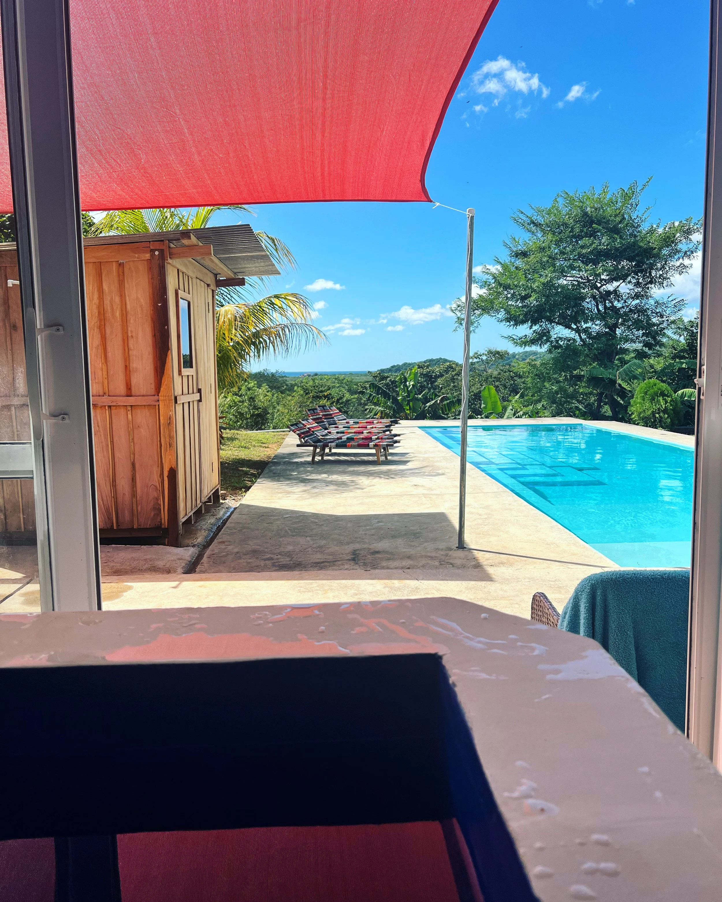 View of a swimming pool outdoors with lounge chairs, green trees, and a blue sky with some clouds, partly covered by a large red shade sail.
