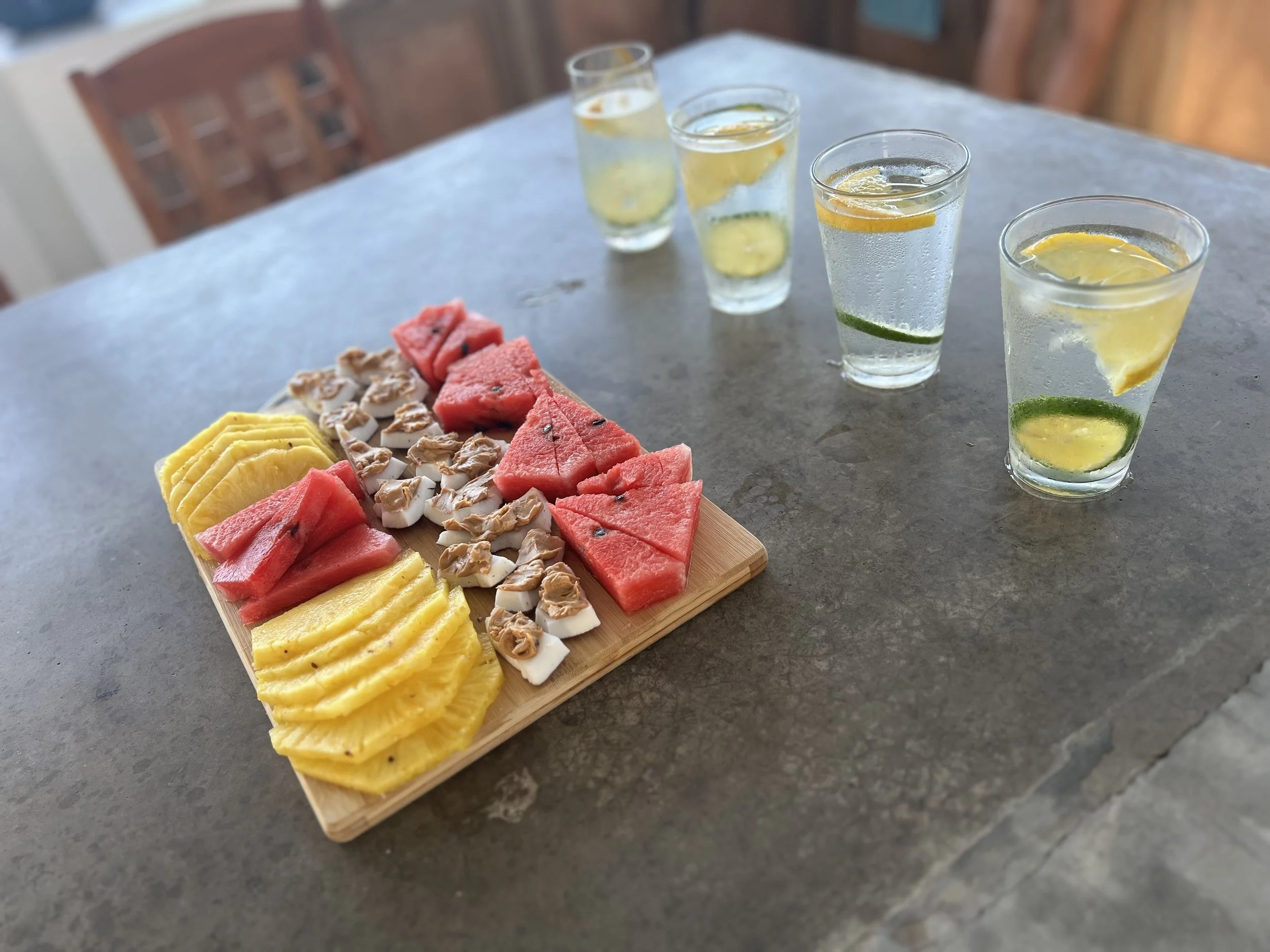 A wooden tray filled with slices of fresh watermelon, pineapple, and coconut topped with walnuts, on a gray stone table with four glasses of lemon and lime infused water.