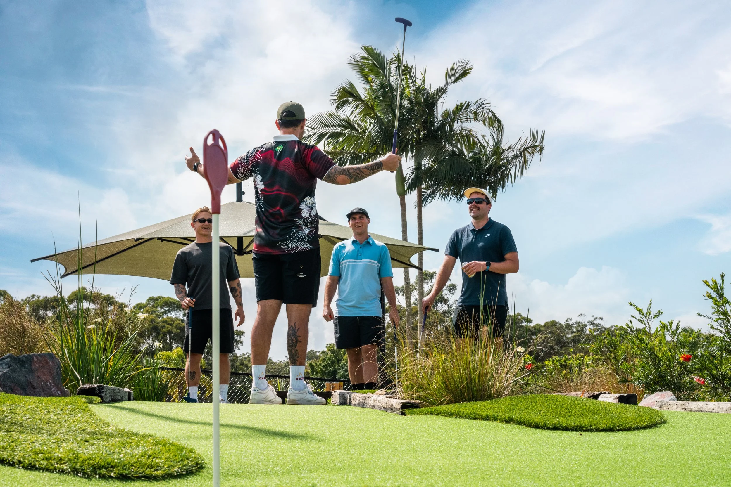 Four men are on a mini-golf course, with one holding a golf club and the others watching, under a partly cloudy sky with palm trees in the background.