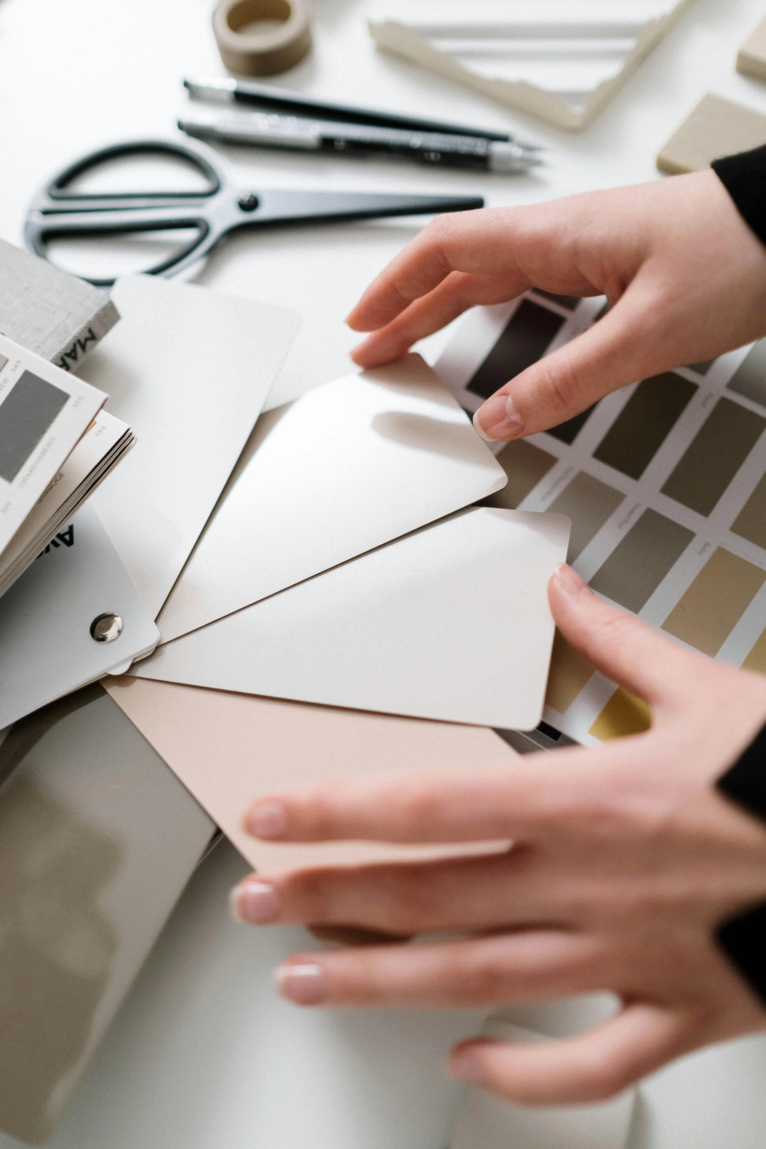 Person holding color swatches or paint samples amidst design tools like scissors, pens, and color guides on a white table.