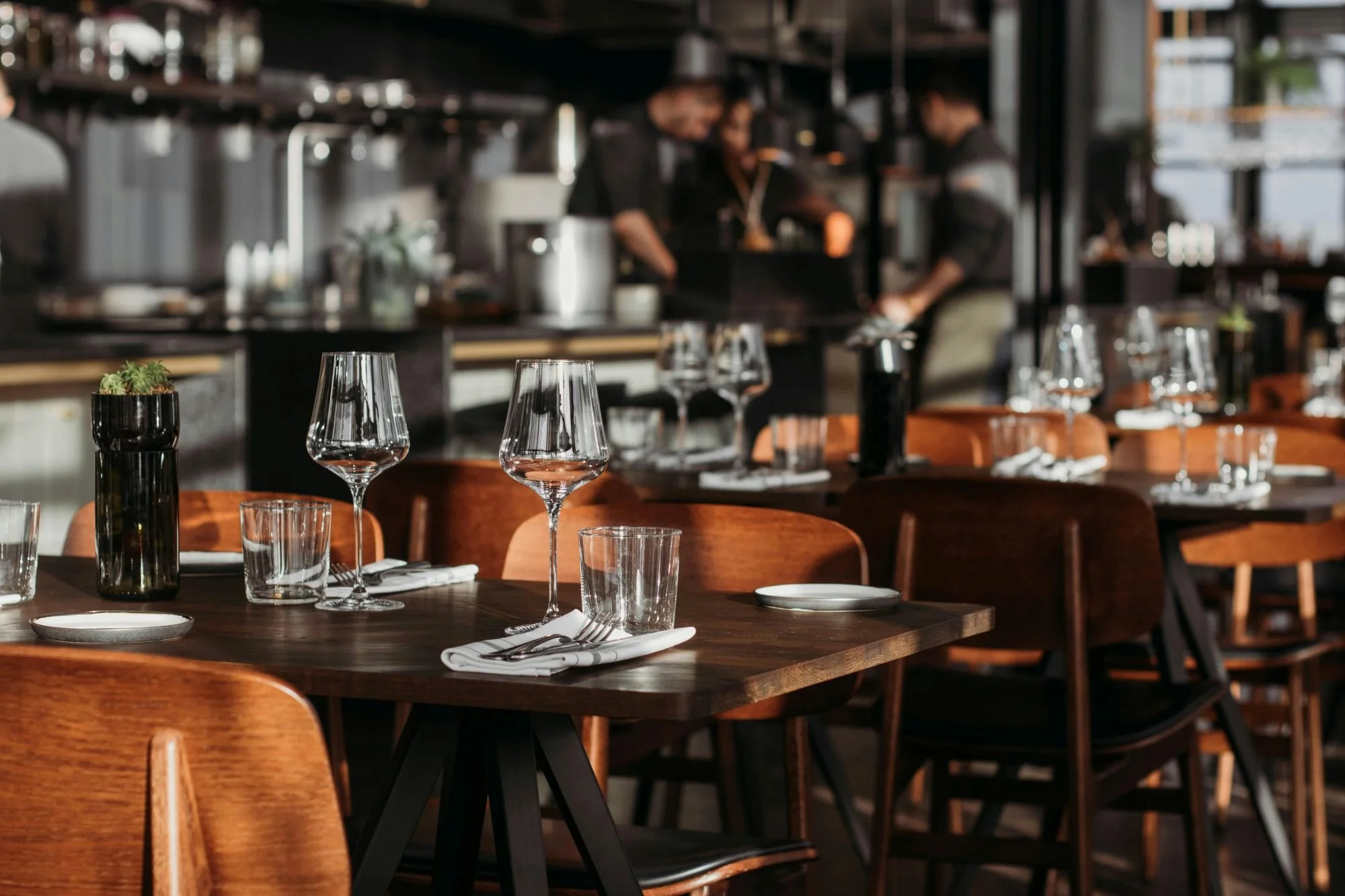 Hospitality interior design. Restaurants, bar and cafes. Empty restaurant tables with wine glasses, forks, white napkins, small plates, and a small green plant in a black vase, inside a modern dining area with wooden chairs.